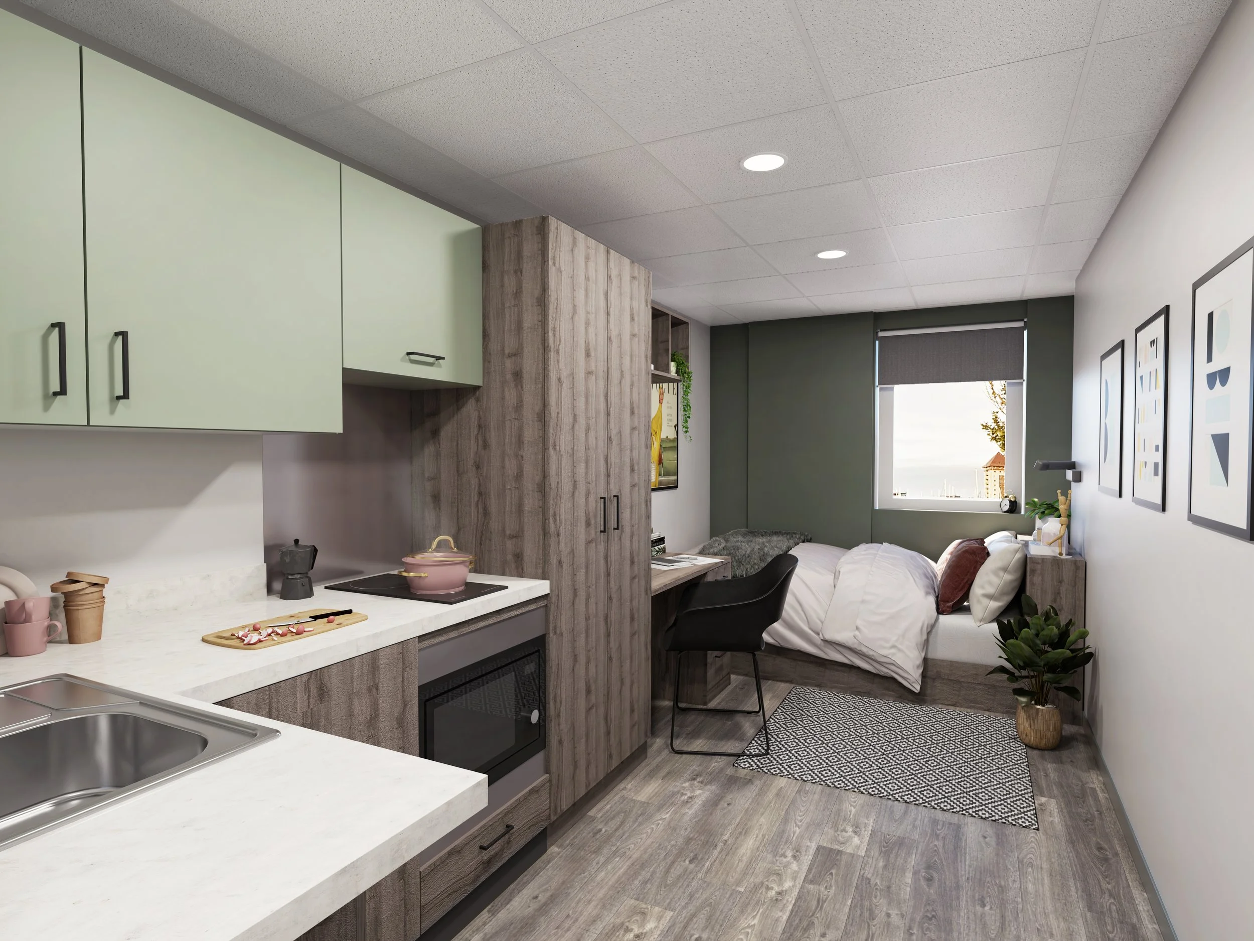 Small modern studio apartment interior with kitchen area, bed, and desk. The kitchen features pale green cabinets, white countertops, and an induction stove. The sleeping area includes a single bed with white bedding, a desk with a chair, and wall ar