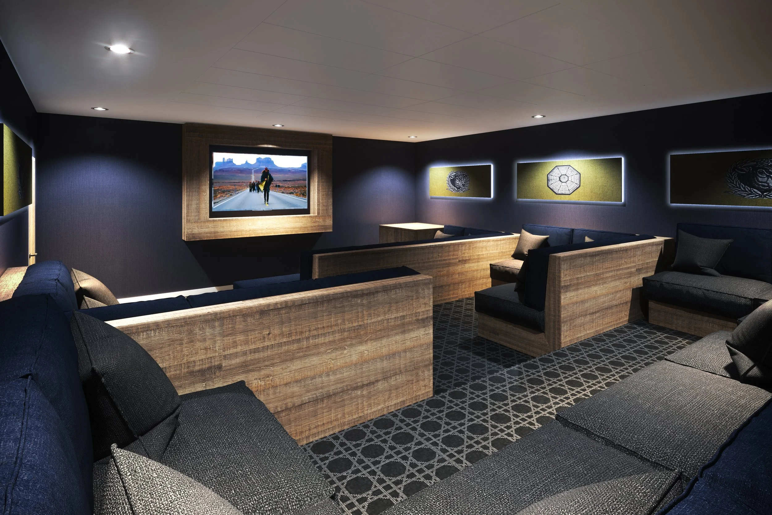 Interior of a modern home theater with several dark-colored, fabric-covered couches and wooden partitions. A large flat-screen TV mounted on the wall displays an outdoor scene. The room has patterned carpeting and decorative wall panels with subtle l