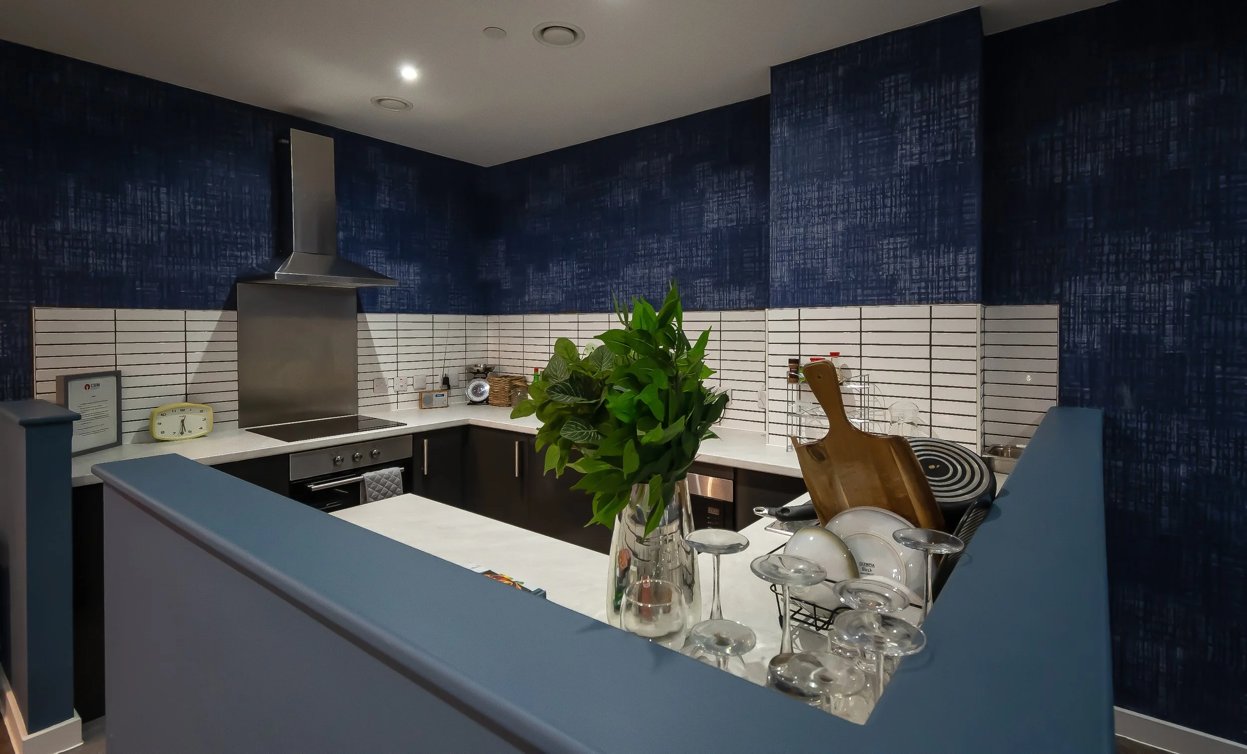 Modern kitchen with blue walls, white countertops, stainless steel range hood, and backsplash. Features plants, a clock, and dishware on counter.