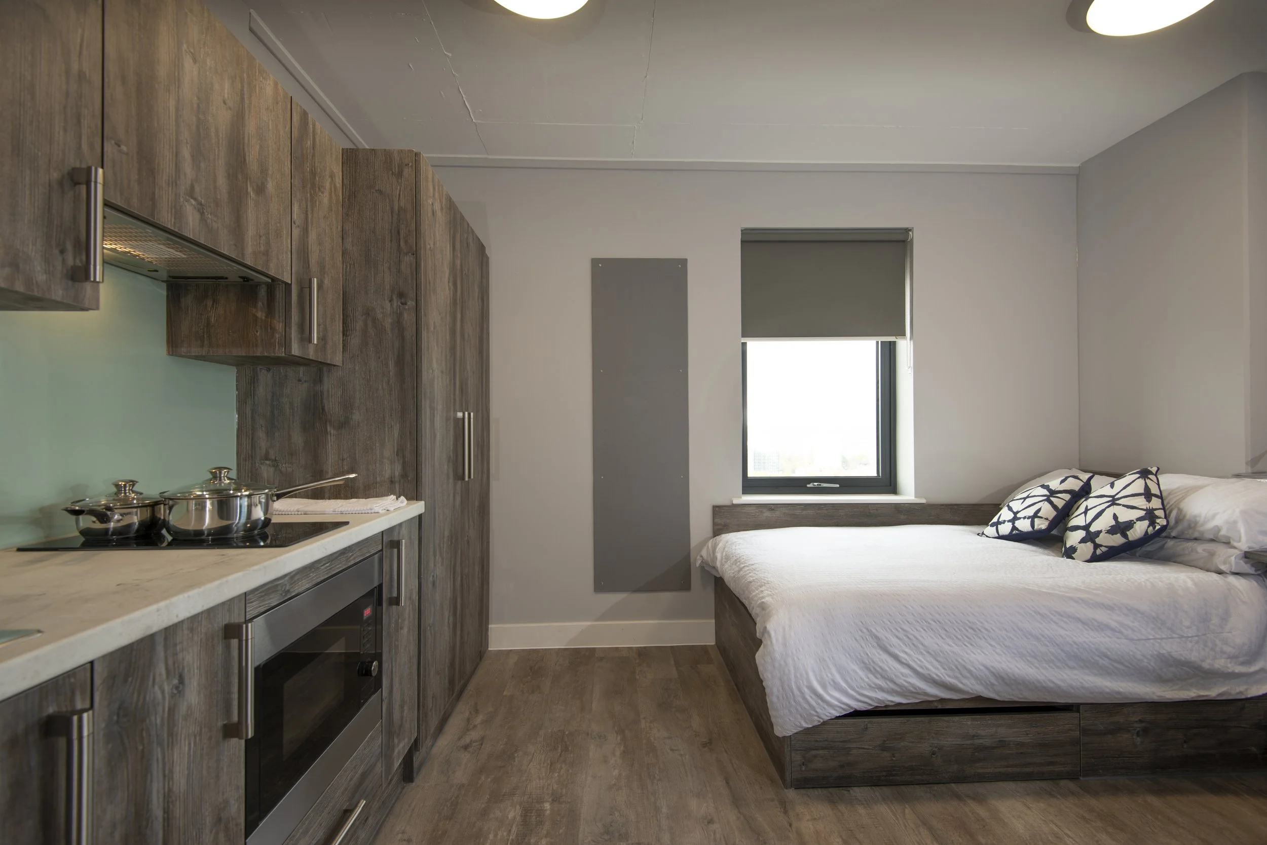 Modern studio apartment interior with a compact kitchen and a bed. The kitchen features wood cabinets, a stovetop, and pots. The bed has white bedding and patterned pillows. A window with a gray blind is present.