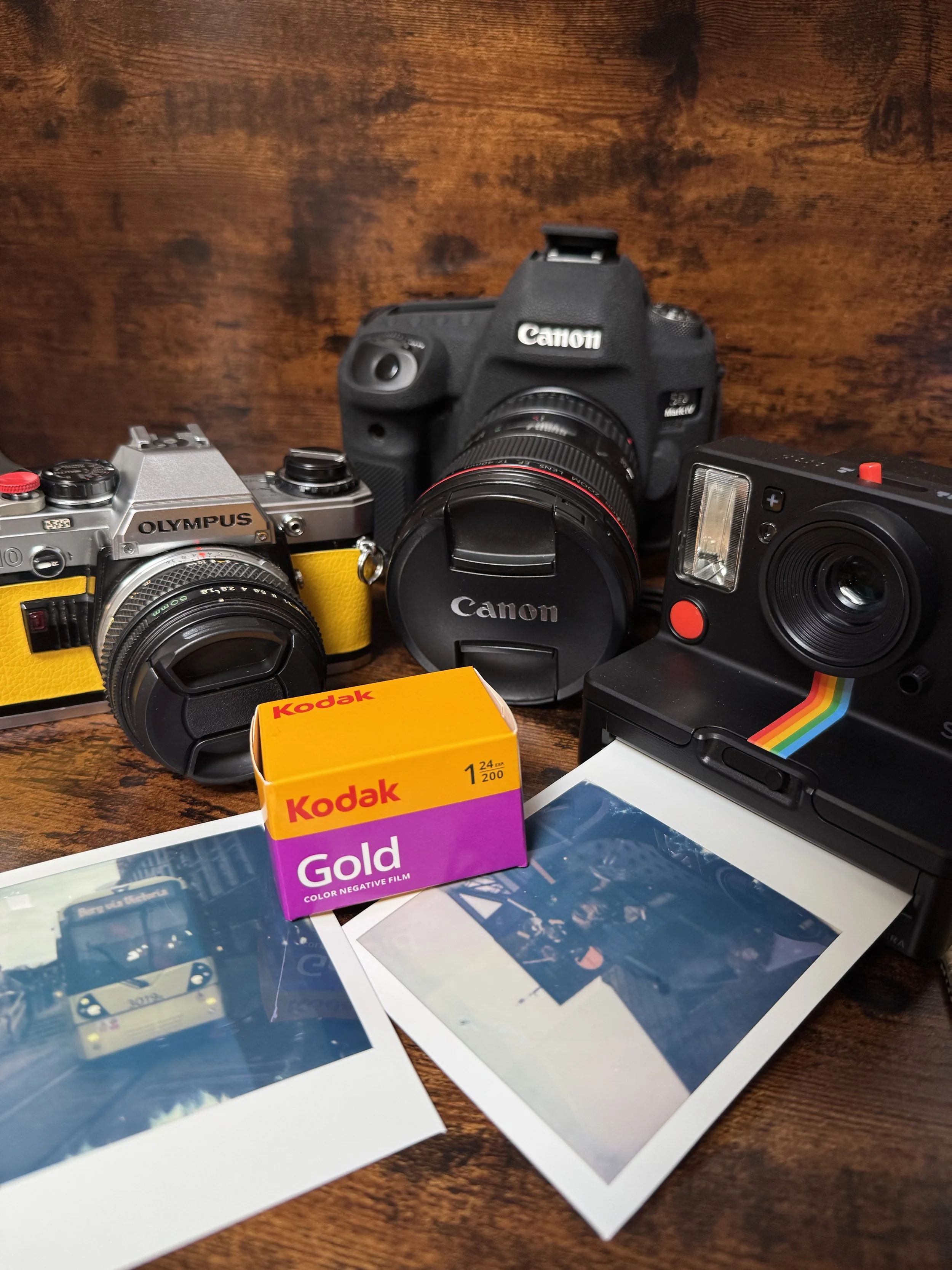 A collection of vintage and modern cameras, a roll of Kodak Gold film, and printed photographs on a wooden surface.