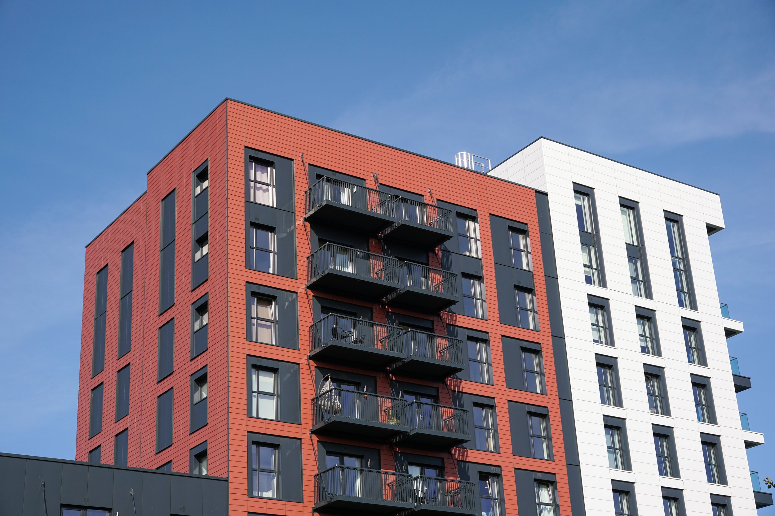 Modern multi-story apartment building with red and white exterior, black balconies, and large windows against a blue sky.