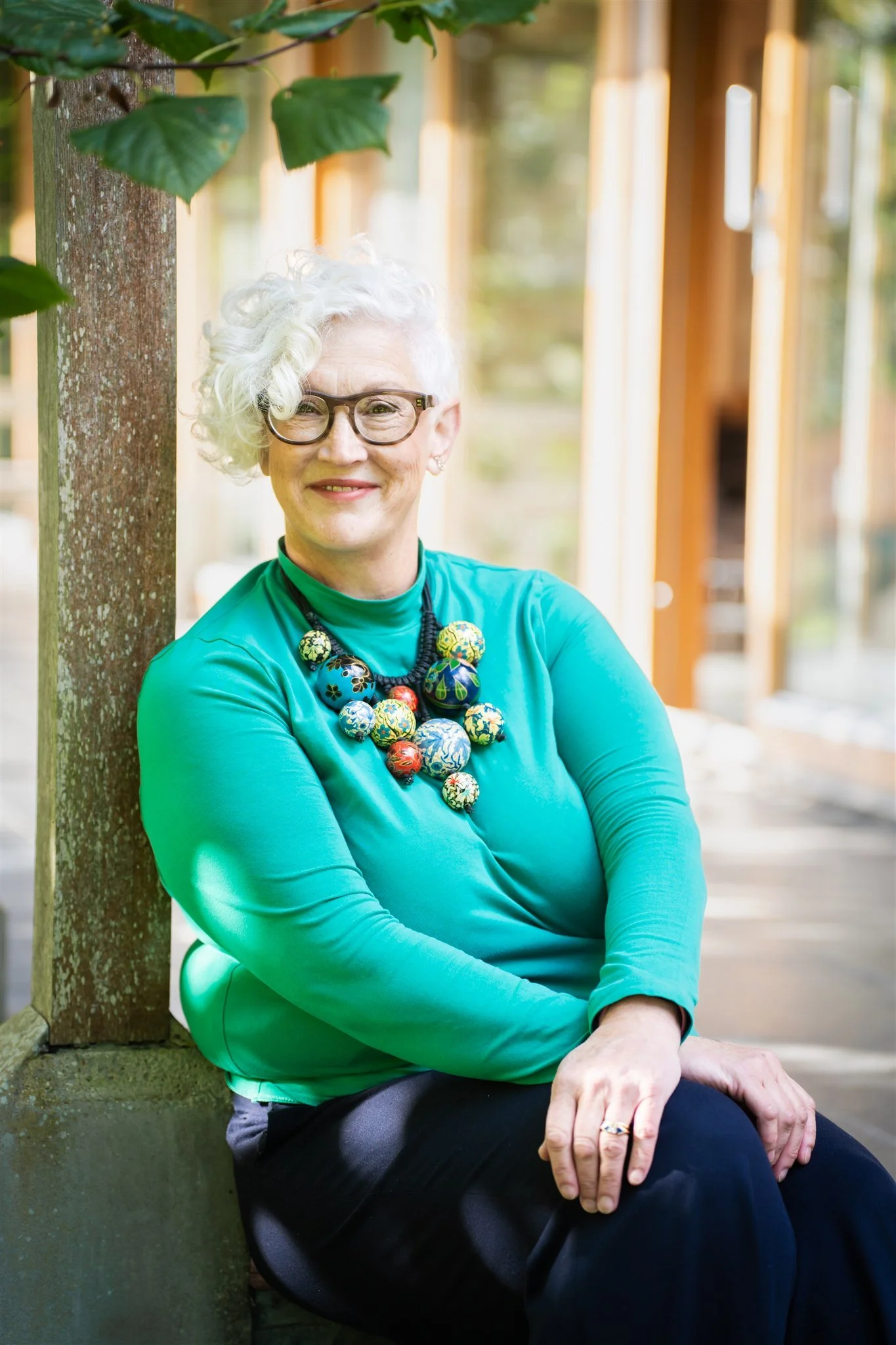 A woman with short, curly white hair and glasses, smiling and sitting outdoors. She is wearing a bright turquoise top and a large colorful beaded necklace. She's sitting against a wooden structure with greenery around her.