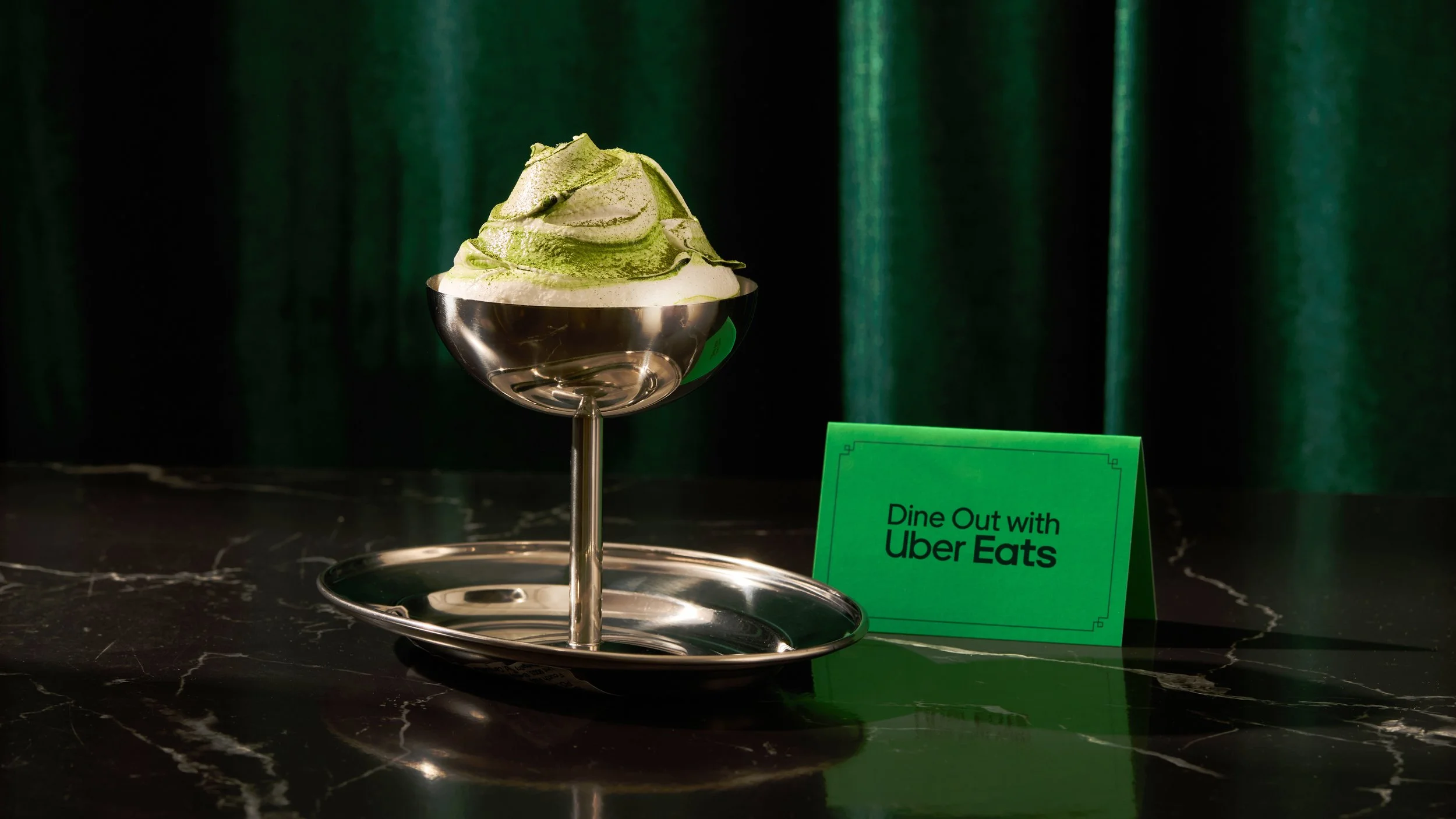 A serving of matcha green tea ice cream topped with powdered green tea in a shiny silver bowl on a black marble surface, with a green sign that reads 'Dine Out with Uber Eats' in the background.