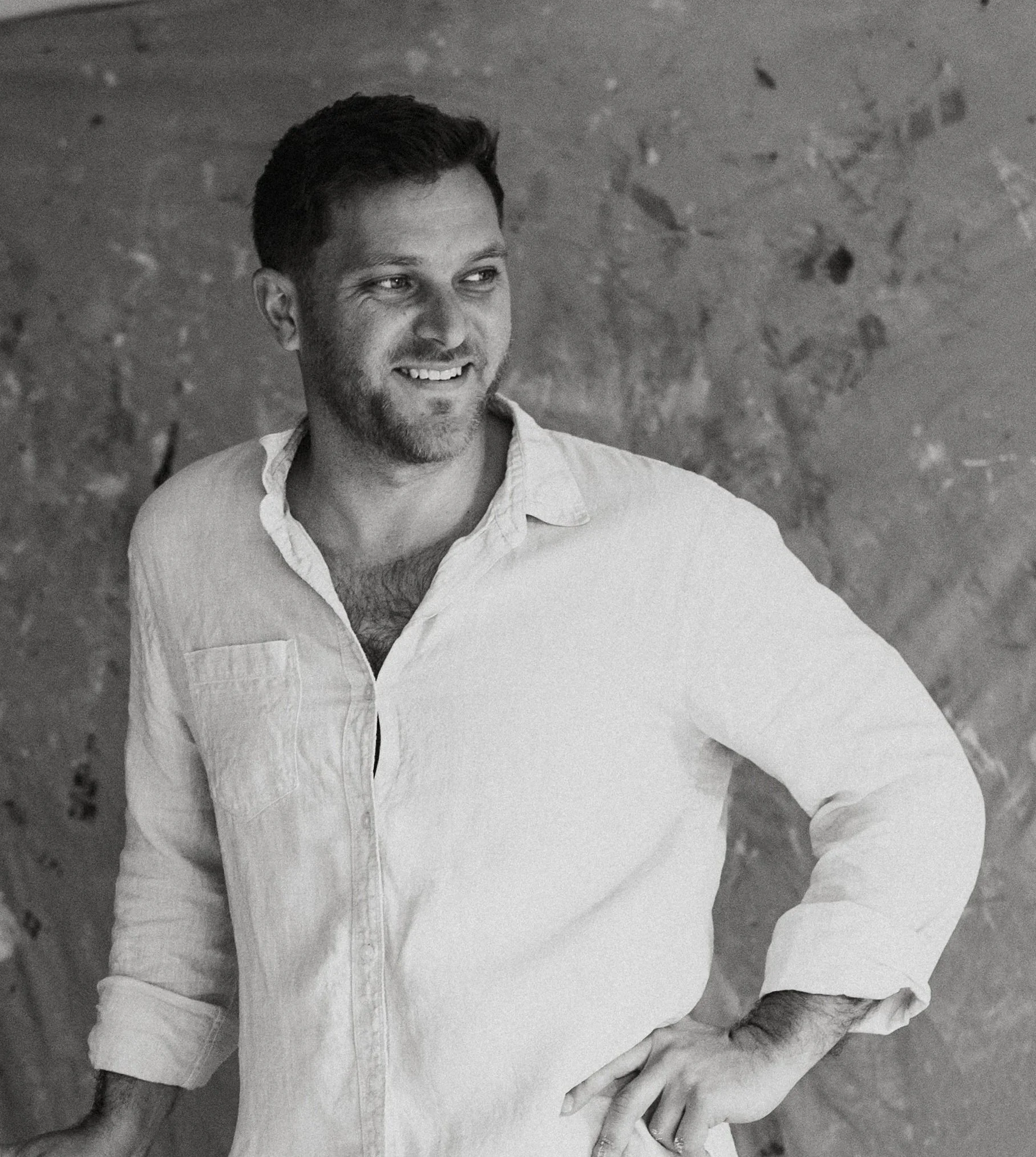 Black and white photograph of a smiling man with short dark hair, beard, and mustache, wearing a light-colored, partially unbuttoned shirt, standing against a textured wall with what appears to be holes or marks.