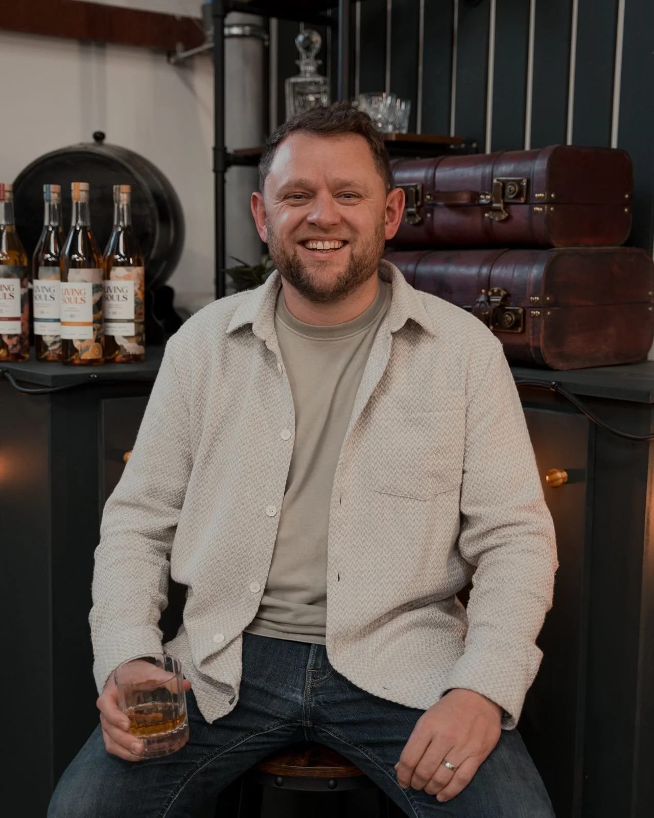 Man sitting with a glass of whiskey, bottles labeled 'Living Souls' behind him, vintage suitcases and glassware in the background.