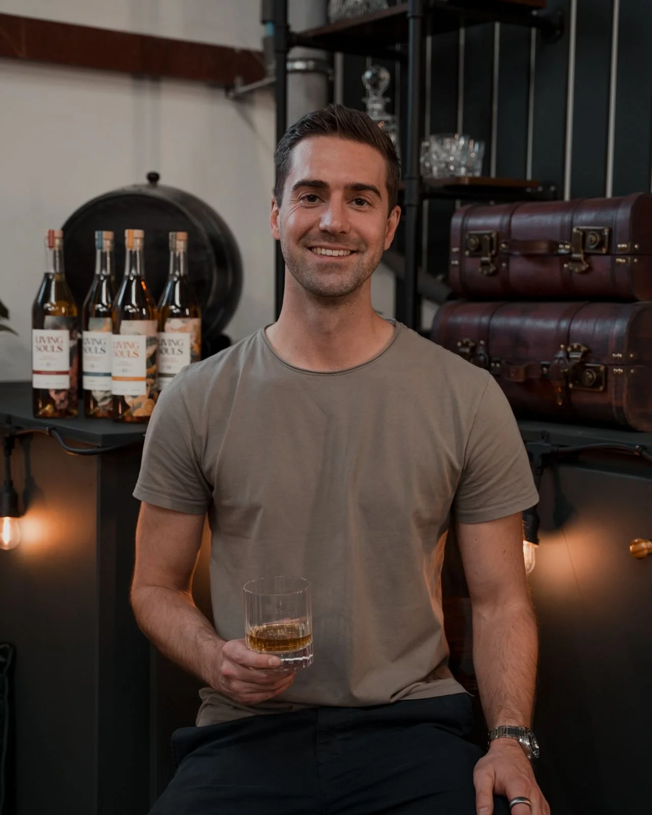 Man holding a drink seated in a bar with four bottles of 'Living Souls' on a table in the background.