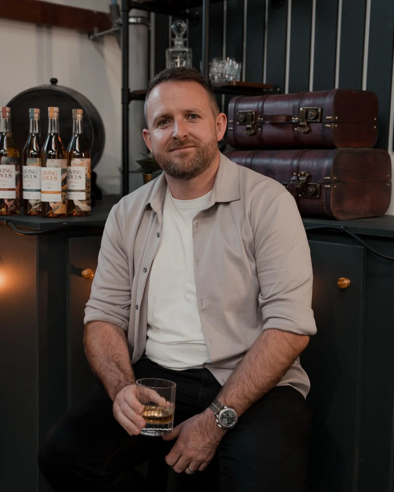 Man sitting with a glass of whiskey, wine bottles in the background, vintage suitcases beside him, wearing a causal outfit and watch.