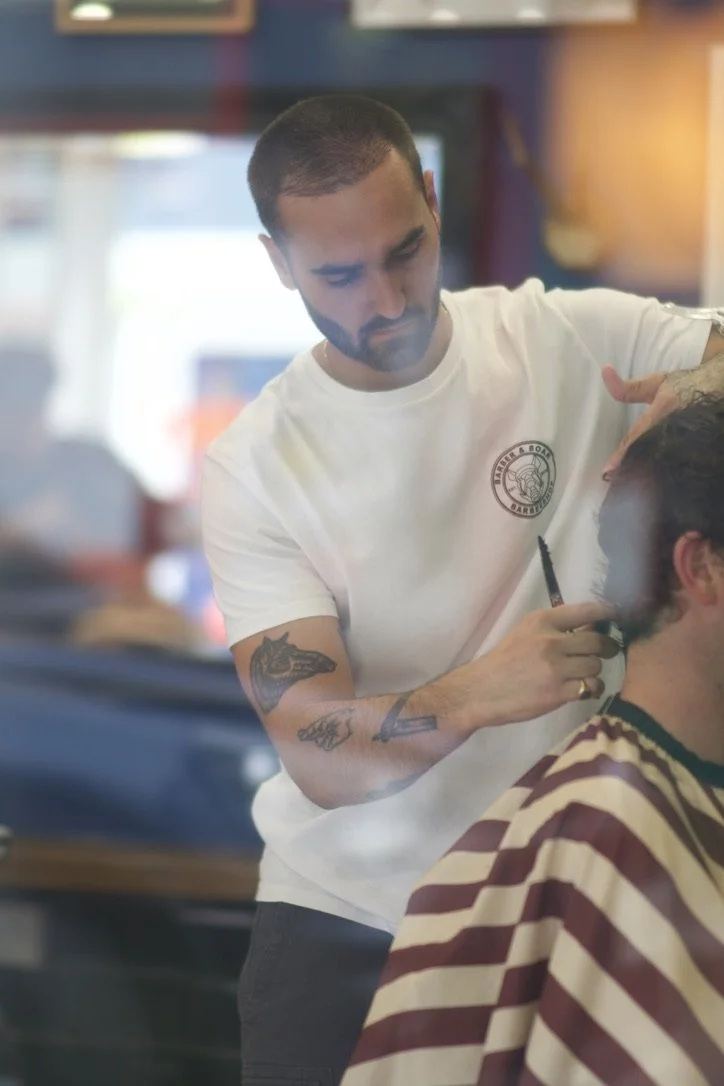 A barber with tattoos on his arms giving a haircut to a customer in a barbershop.