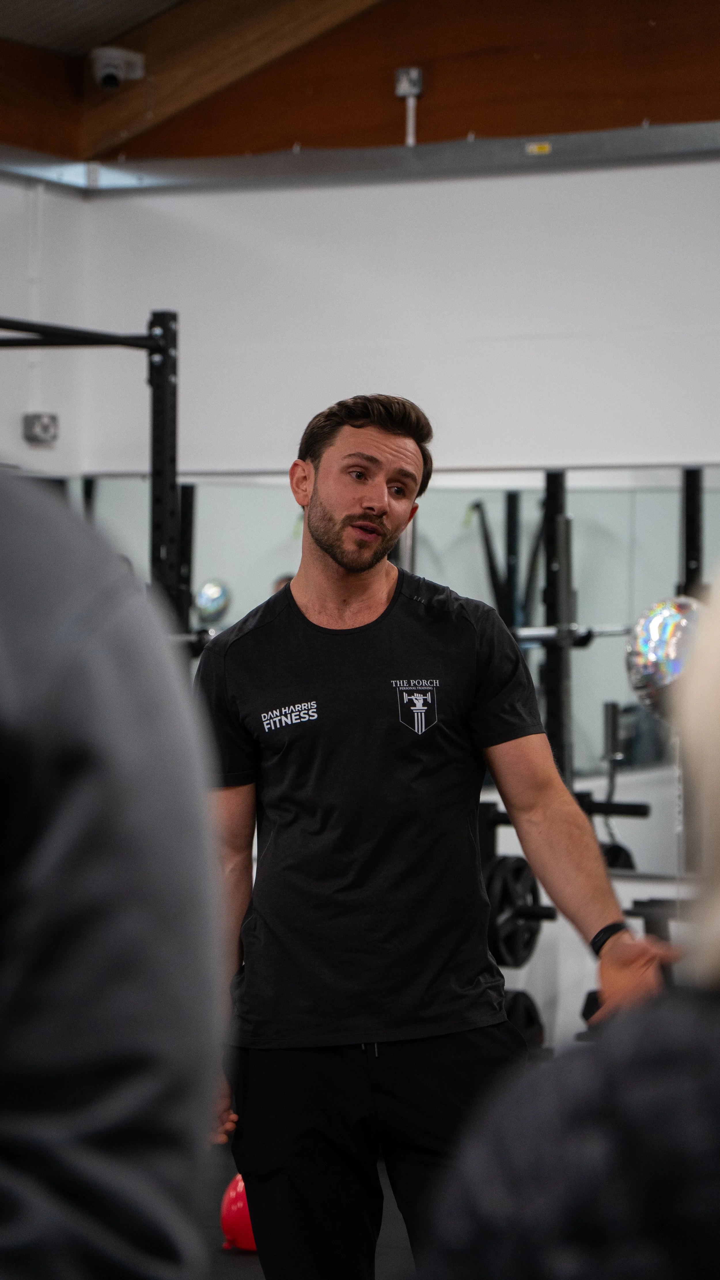 A fitness trainer standing in a gym, talking to a group. He is wearing a black T-shirt with the words 'DAN HARRIS FITNESS' and a logo that says 'THE PORCH'.