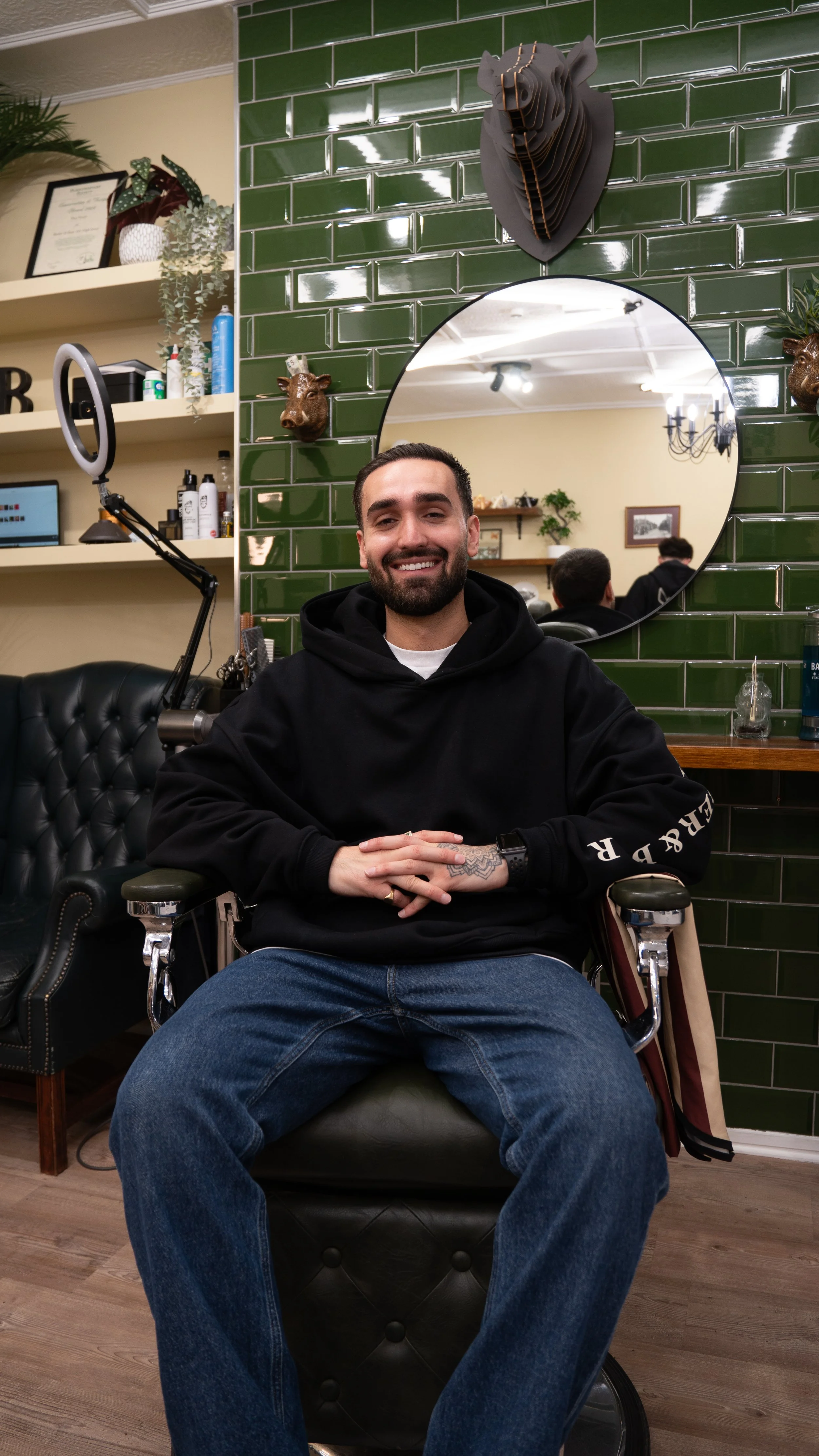 A smiling man with a beard, wearing a black hoodie and jeans, sitting in a barber chair in a salon or barbershop with green tiled wall, mirrors, and decorative animal heads.