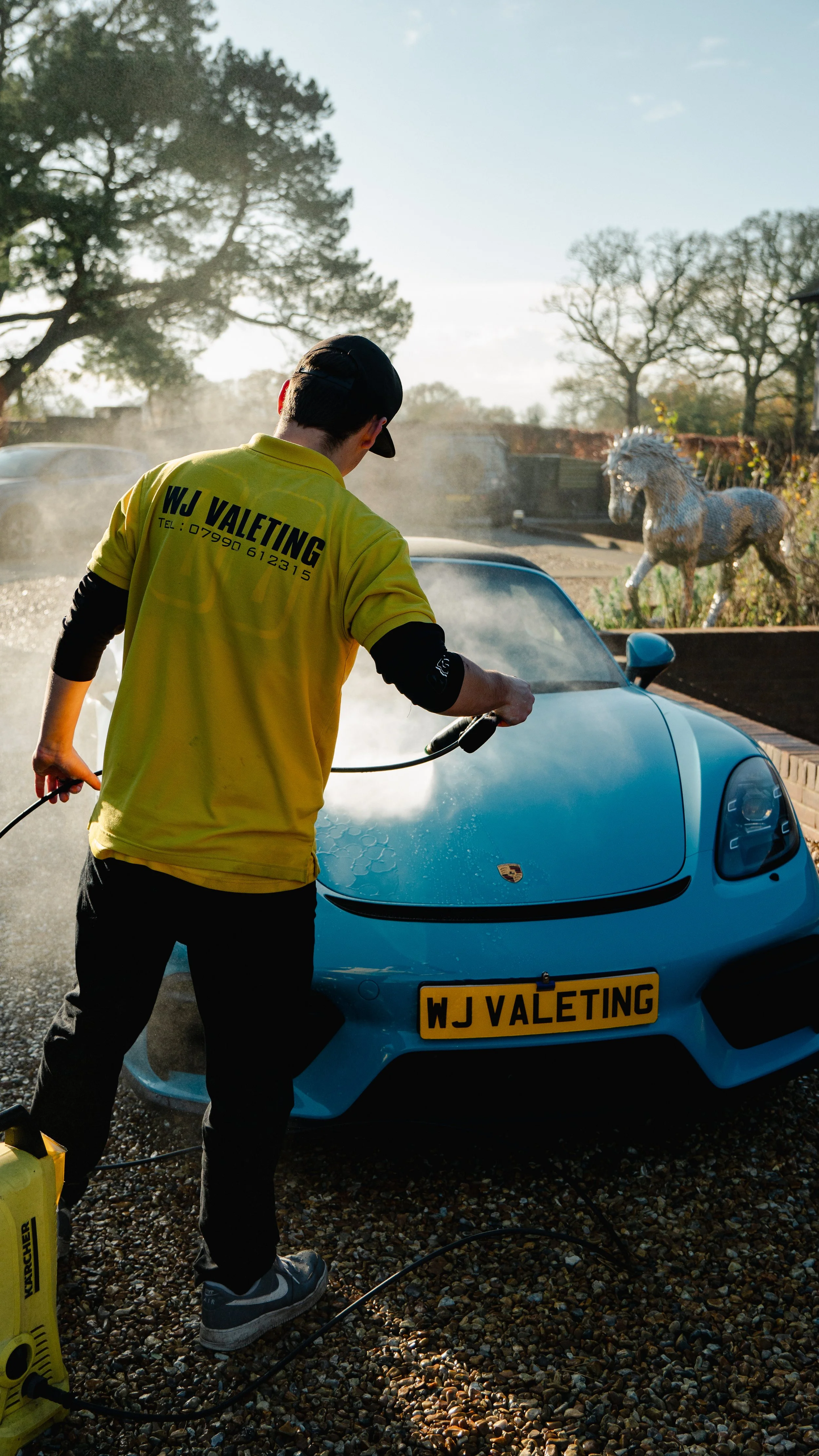 A person washing a blue Porsche sports car outdoors with a pressure washer, wearing a yellow shirt with 'WJ Valeting' on the back, and a black cap. There are trees and a decorative horse statue in the background.