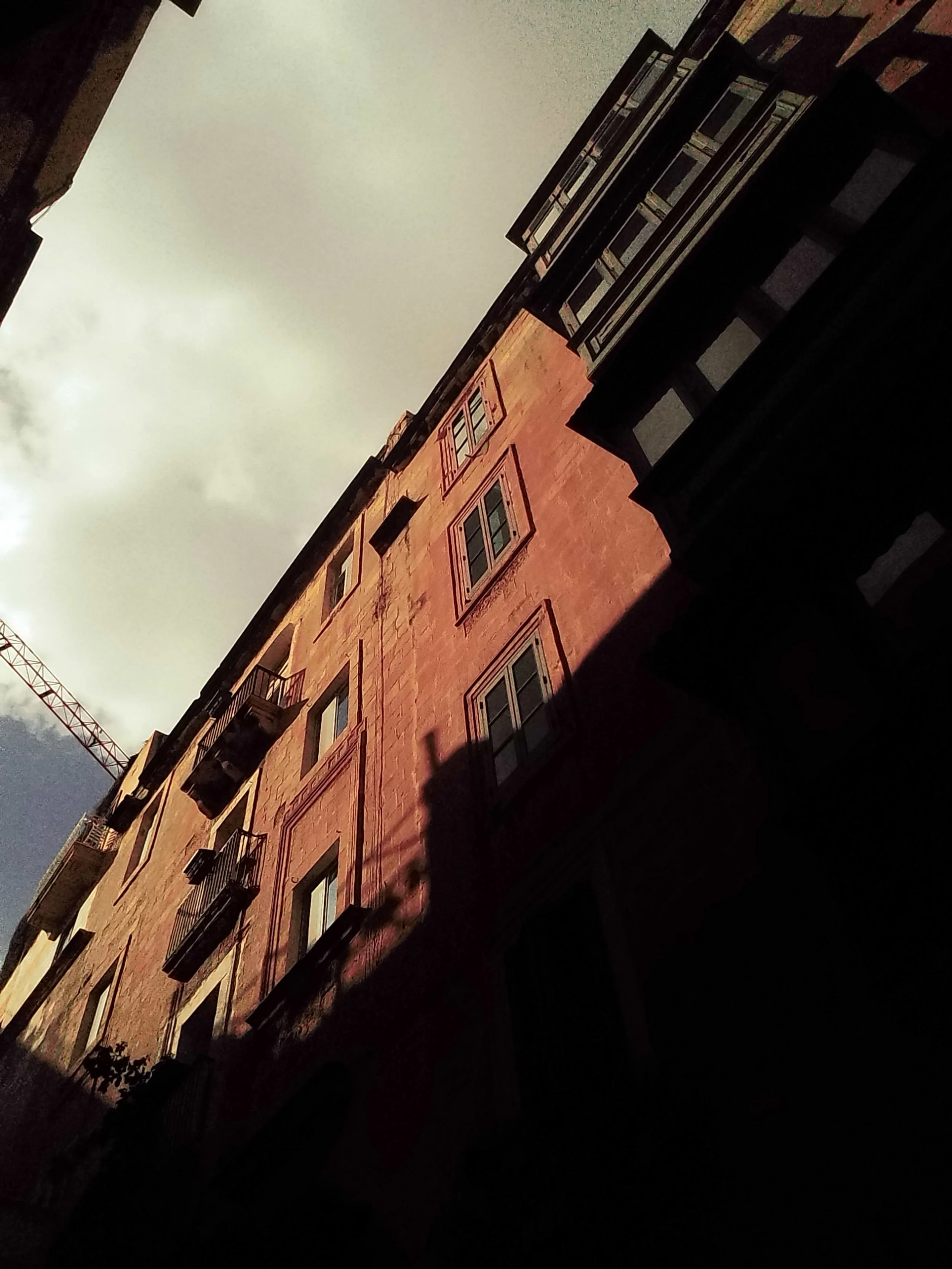 View of an old pink building with balconies and windows, with shadows cast on it, under a cloudy sky.