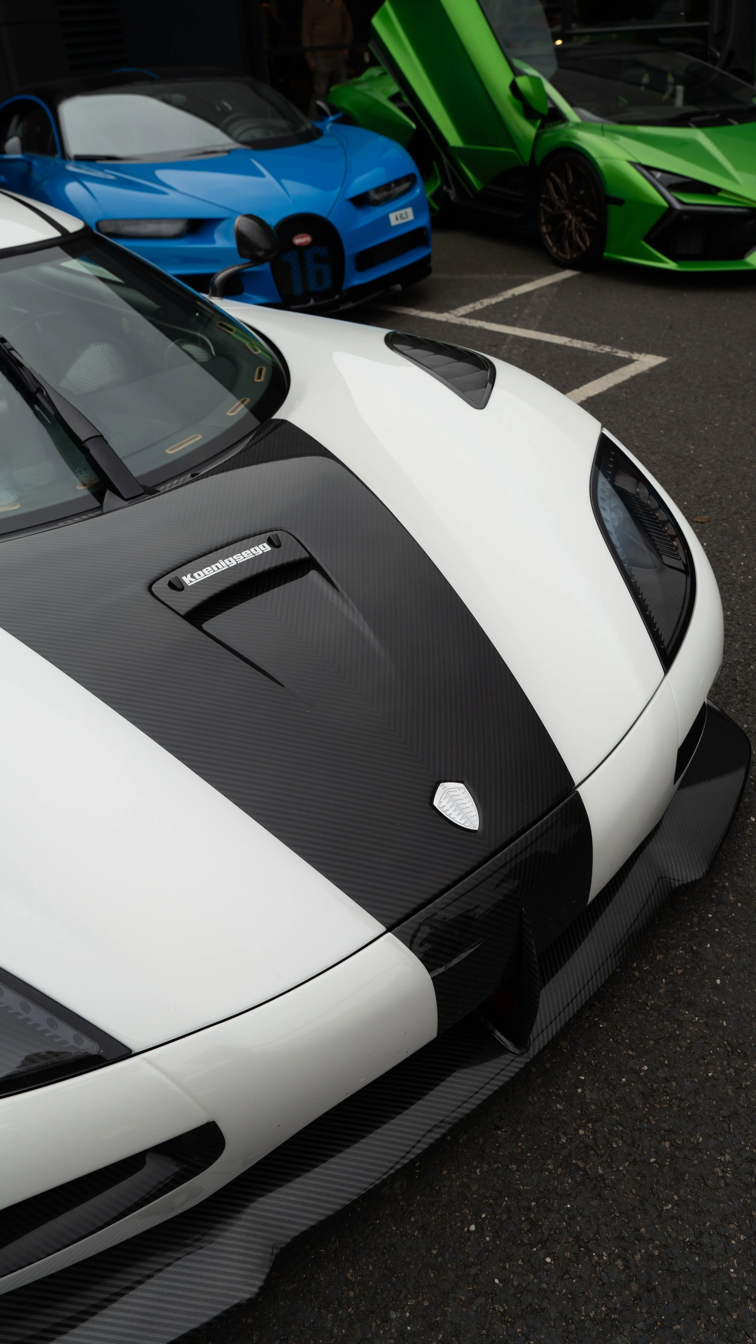Close-up of a white and black Koenigsegg sports car with carbon fiber accents, parked among other luxury vehicles including a blue sports car and a green Lamborghini.