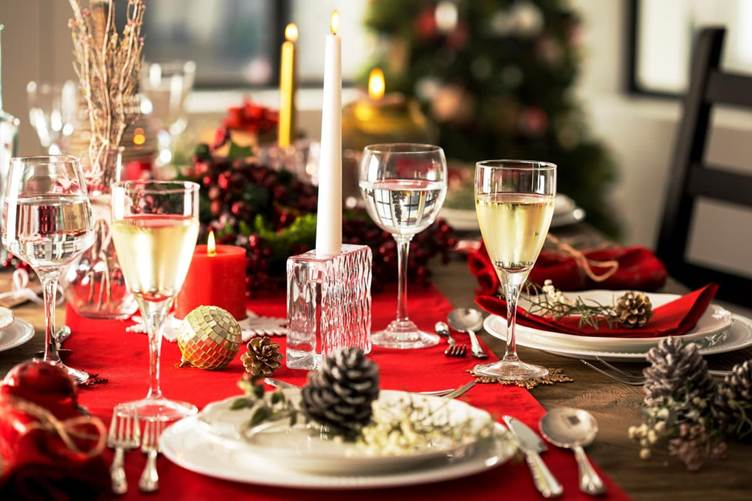 Festive holiday table setting with candles, wine glasses, pinecones, and Christmas decorations on a red tablecloth.