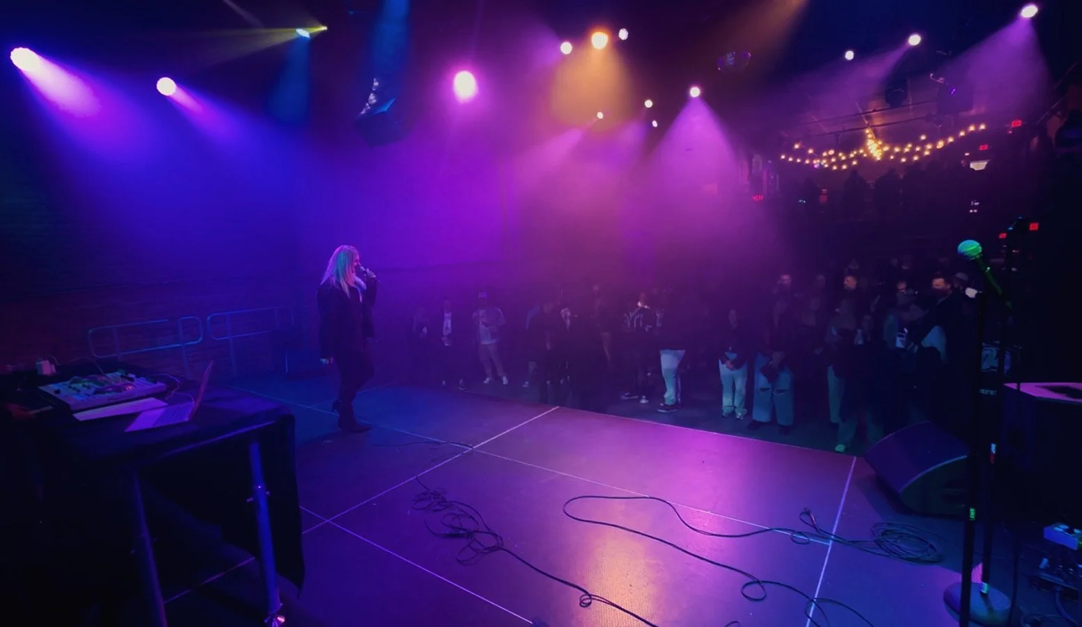 American singer-songwriter Meriele singing on stage with purple and yellow stage lights, an audience watching.