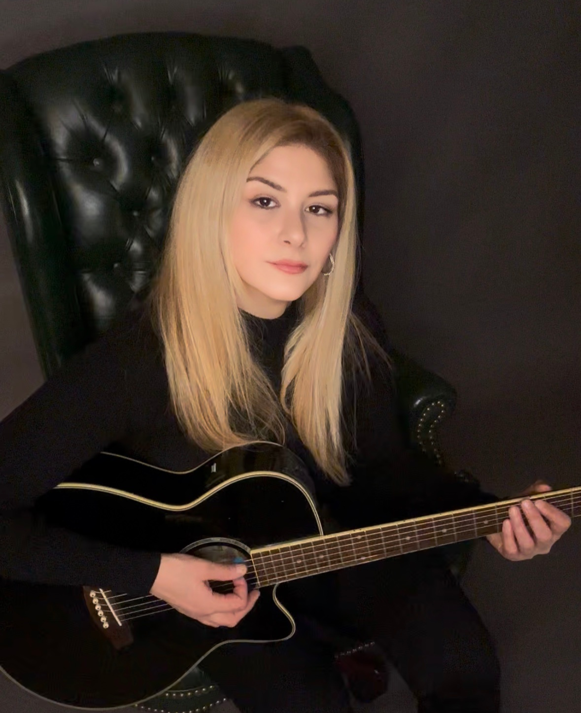 A young woman with long blonde hair sitting on a black leather armchair, playing a black acoustic guitar, against a black background.
