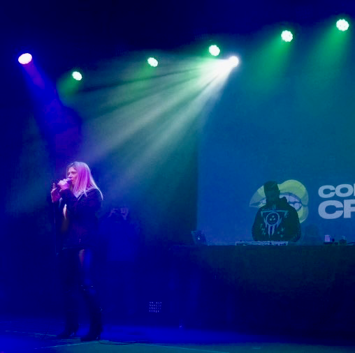 A woman singing into a microphone on a stage illuminated by green and blue stage lights, with a DJ or sound booth in the background featuring a cartoonish character and the words "CO" and "CR".
