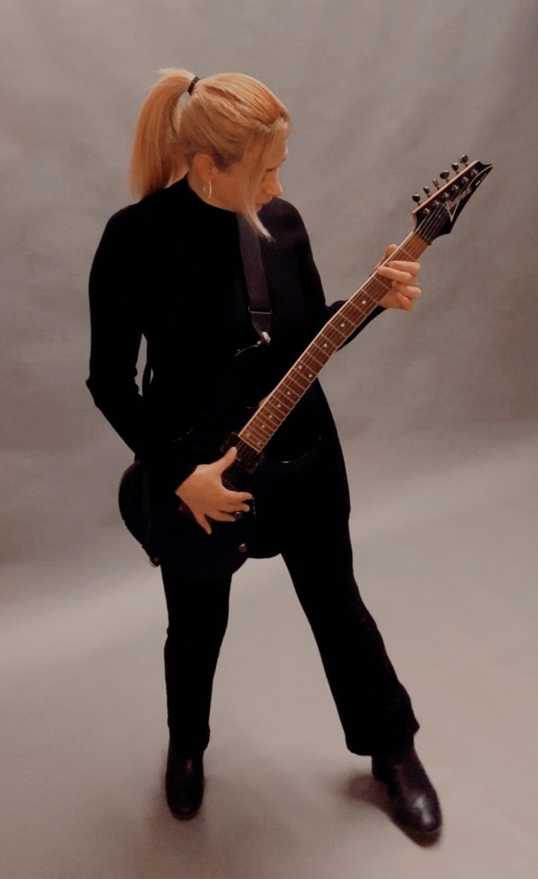 A woman with blonde hair tied back playing an electric guitar, dressed in black, against a plain gray background.