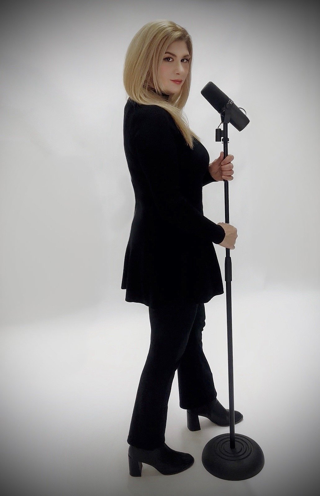 A woman with blonde hair in a black outfit standing sideways, holding a microphone on a stand, against a plain light background.