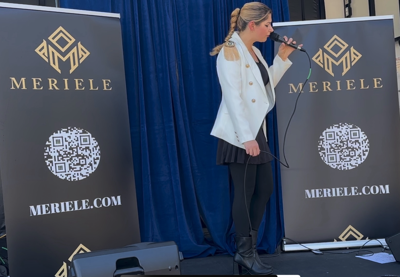 Woman in white blazer and black dress standing on stage, speaking into a microphone at Meriele event, flanked by Meriele banners with QR code and website.