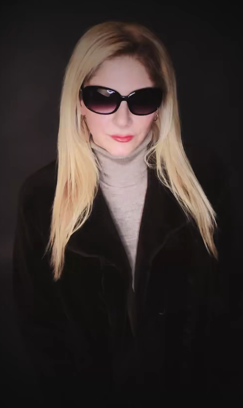 A woman with long blonde hair wearing large black sunglasses, a light gray turtleneck, and a black coat against a dark background.