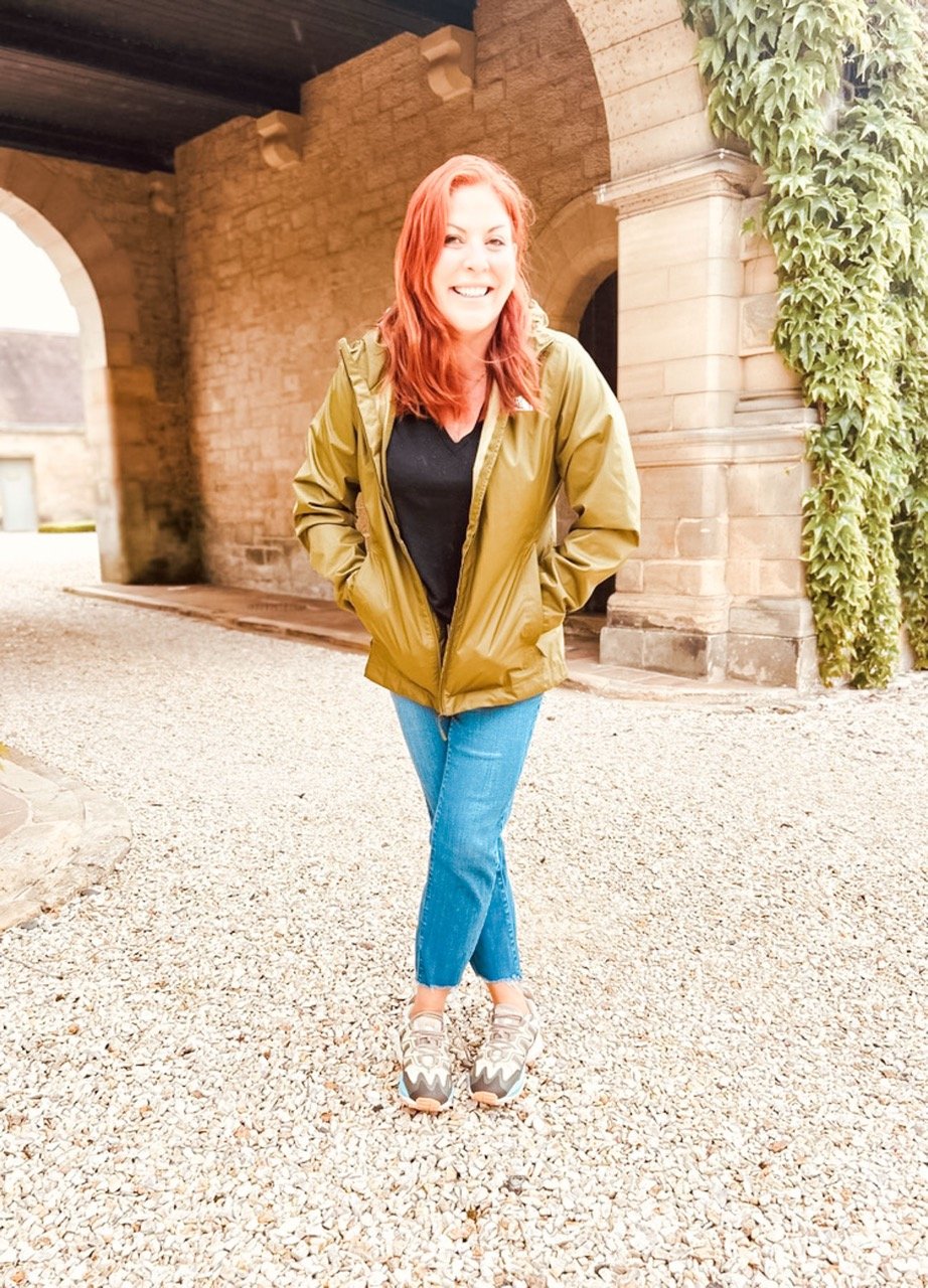 A woman with red hair and a big smile, wearing a green jacket, black shirt, blue jeans, and sneakers, standing outdoors in front of an old brick building with ivy growing on the side.