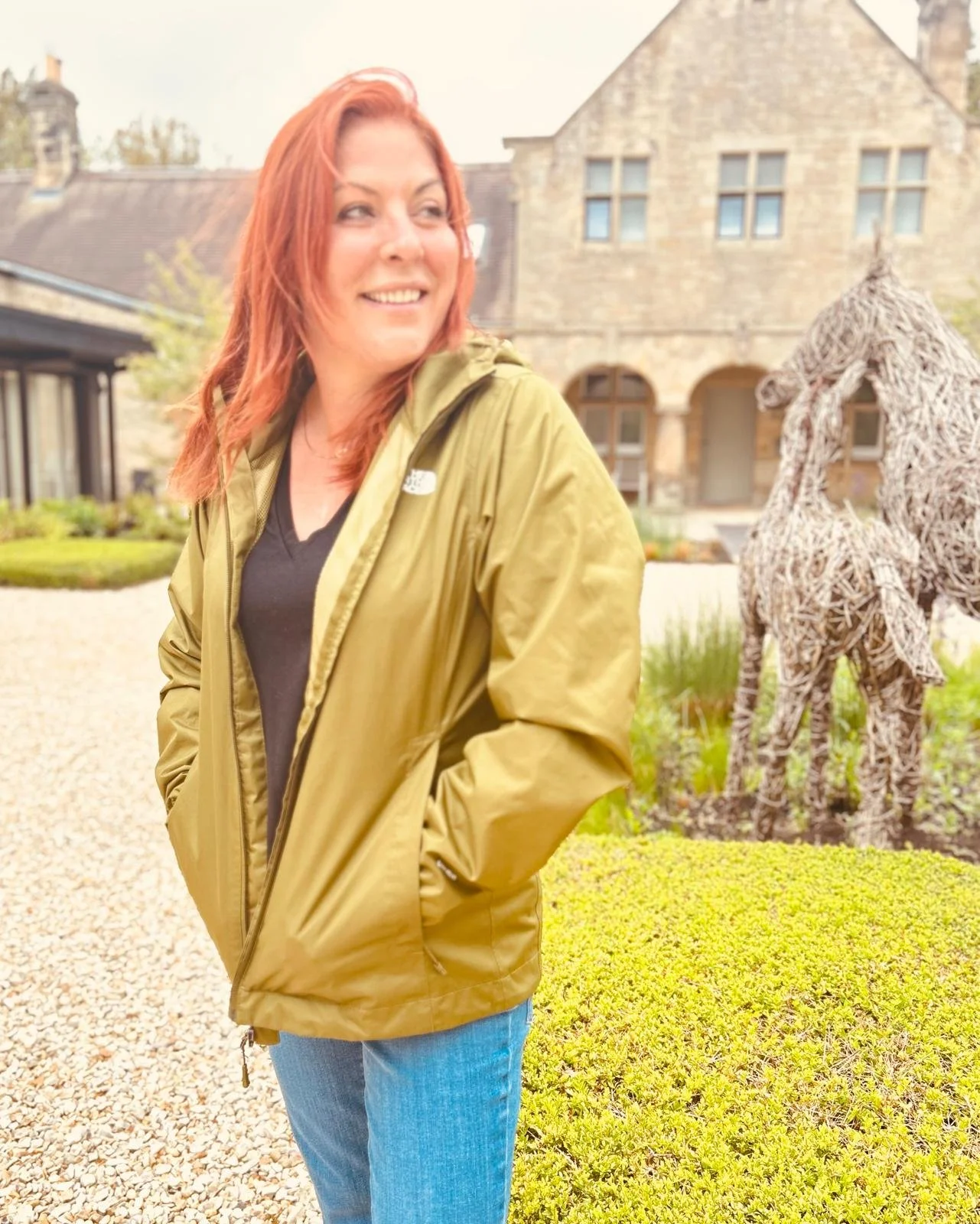 A woman with red hair wearing a green jacket and blue jeans standing outdoors in front of a sculpture of two horses made of woven material, with a large stone house in the background.