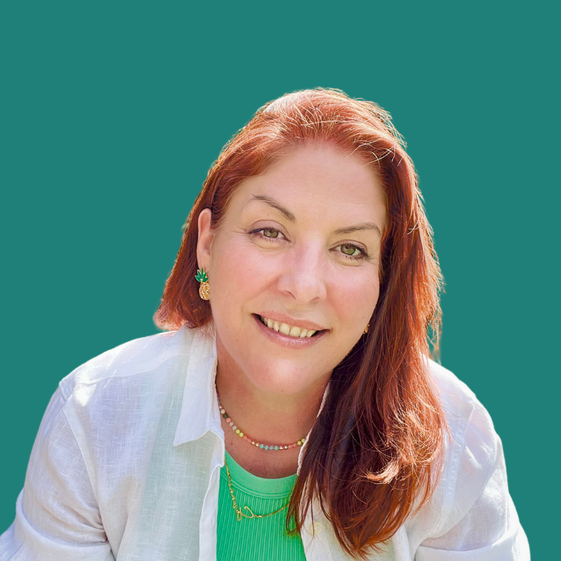 A woman with red hair smiling, wearing a white shirt, green top, colorful jewelry, and pineapple earrings, against a teal background.