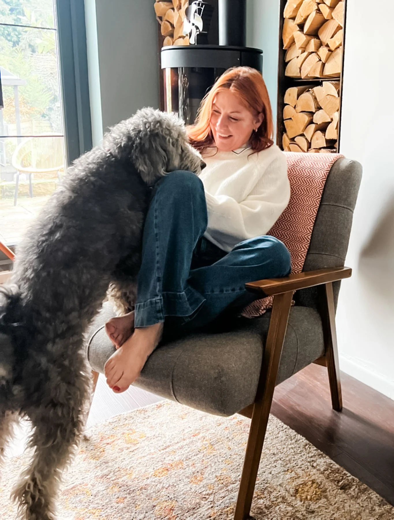 A woman sitting in a gray armchair with her legs crossed, smiling at a large, fluffy dog that is standing on its hind legs with its front paws on her lap. The woman has red hair and is wearing a white sweater and jeans. There is a wood-burning stove and firewood stacked behind her.