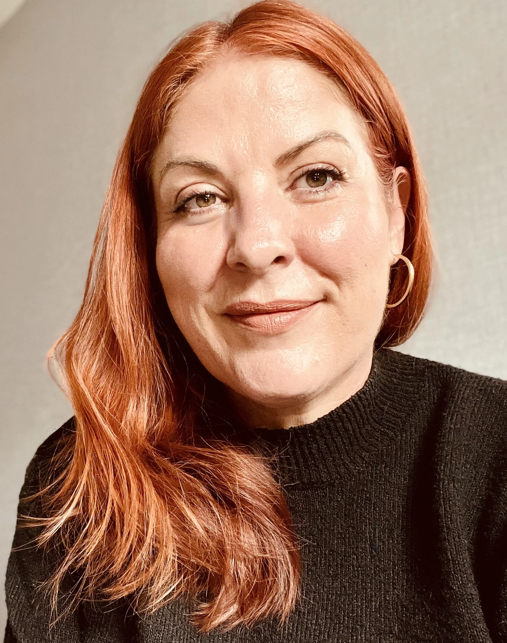 Close-up portrait of a woman with red hair, fair skin, and wearing a black sweater and hoop earrings, smiling softly with a neutral background.