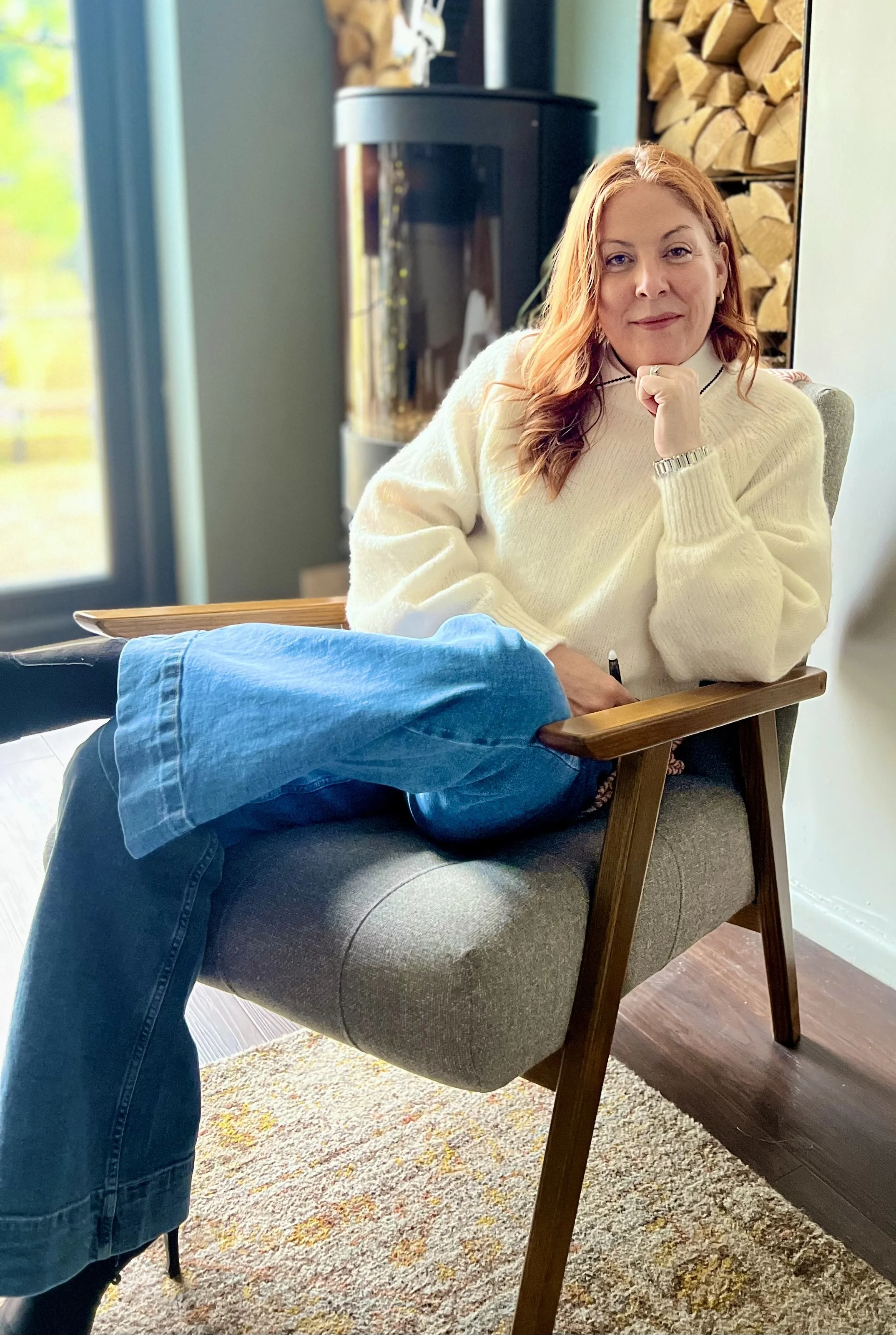 A woman with red hair sitting in a chair at home. She is wearing a cream sweater and has her hand under her chin. There is a wood-burning stove and a pile of firewood in the background.