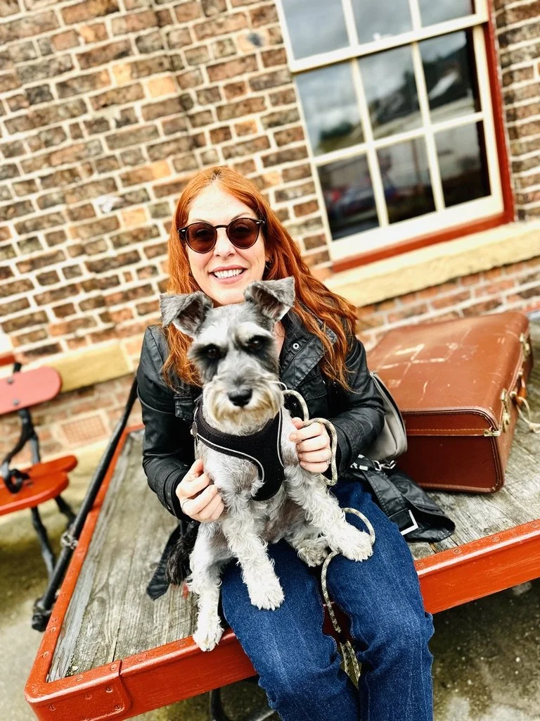 Woman with red hair wearing sunglasses and a leather jacket sitting on a wooden bench outside, holding a small Schnauzer dog in her lap. Brick wall and window in background.
