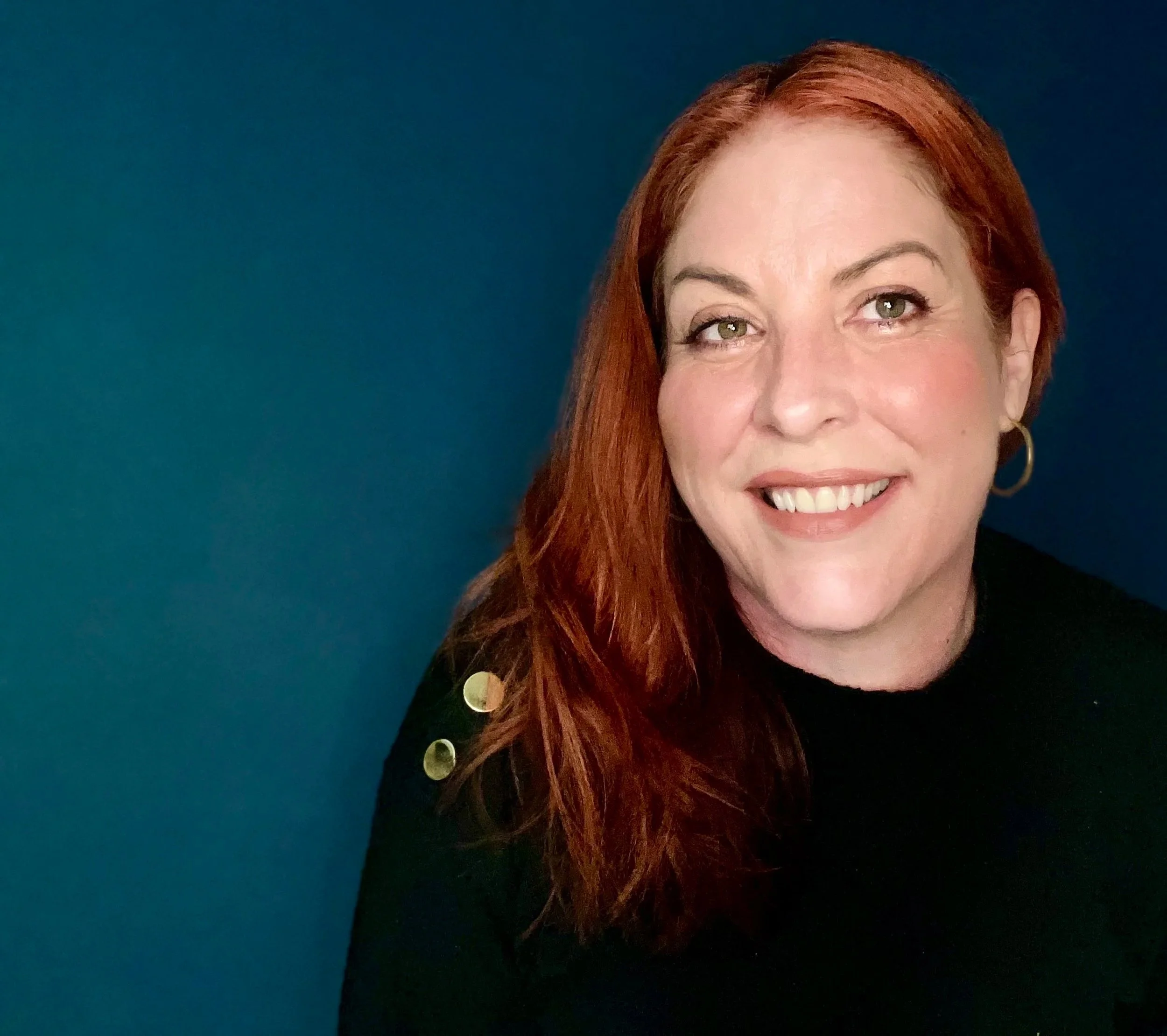 A woman with red hair smiling in front of a blue background, wearing a black top with gold buttons on the shoulder and gold hoop earrings.
