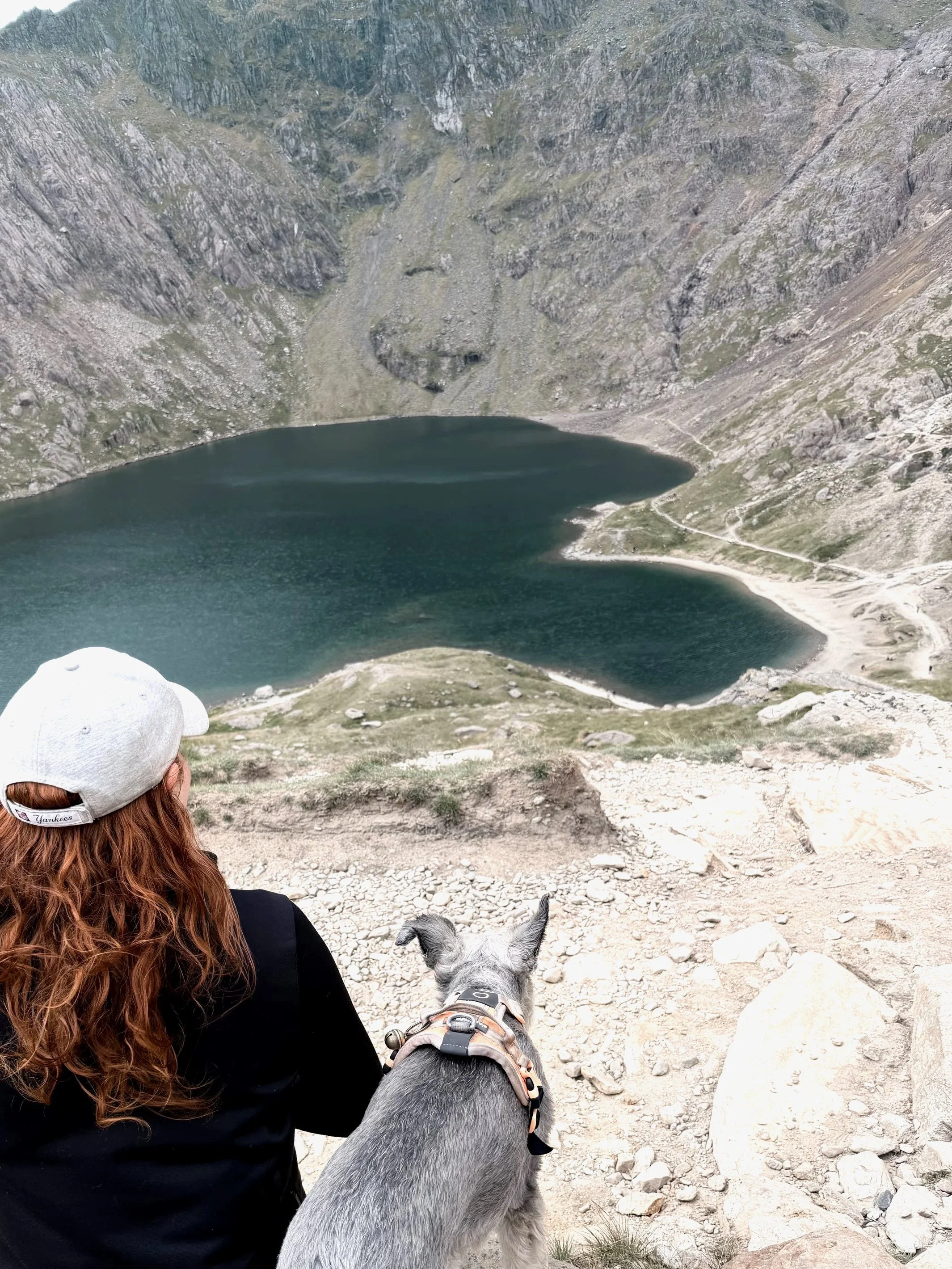 A woman with long curly red hair wearing a white baseball cap and black clothing, sitting outdoors next to a dog with gray fur and a harness, overlooking a mountain lake with rocky slopes and a trail along the shoreline.