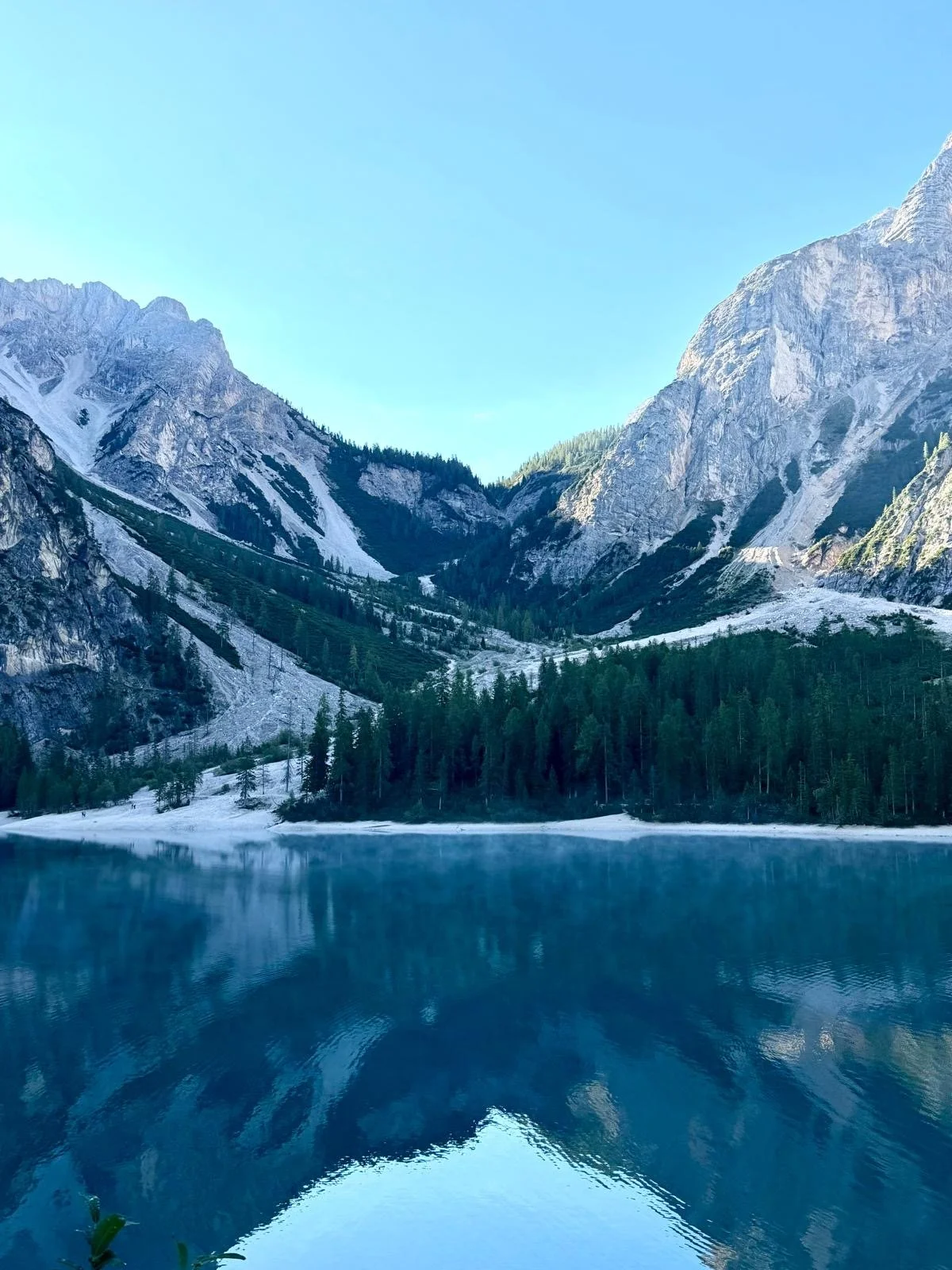 Mountain range with snow patches, a forested hillside, and a reflective lake in the foreground under a clear blue sky.