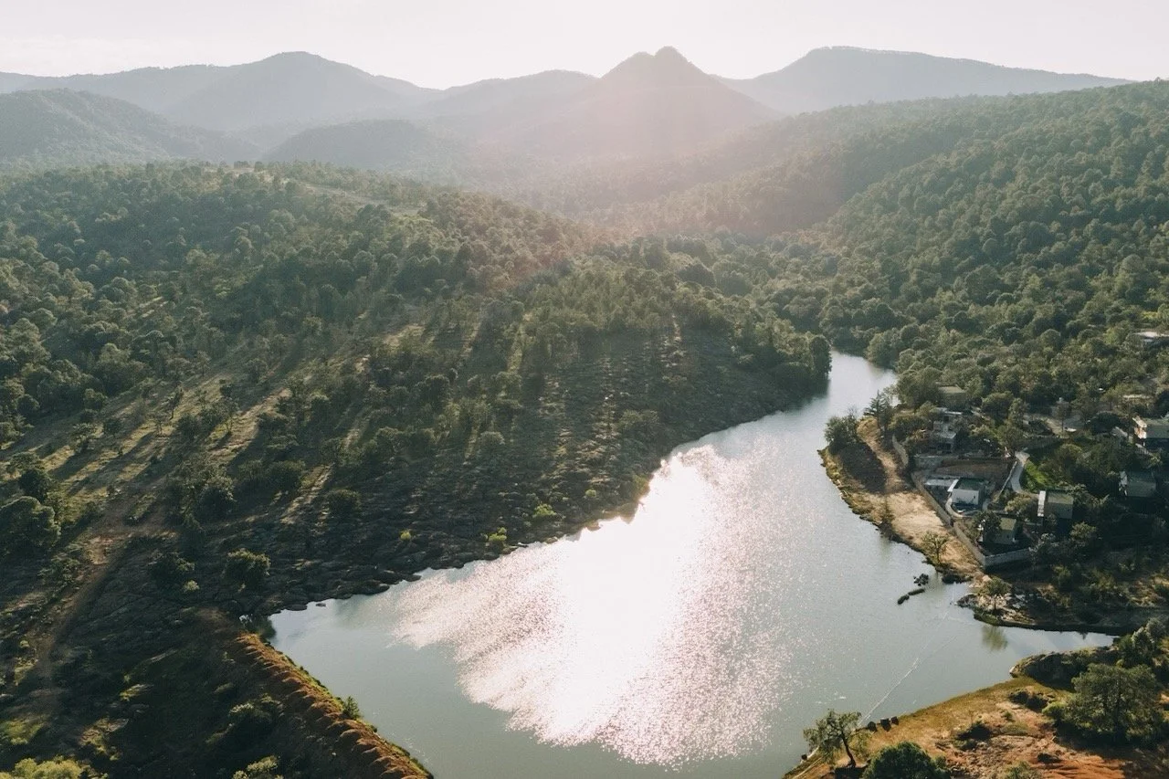 A scenic aerial view of a river winding through a green, mountainous landscape during late afternoon or early evening with the sun reflecting off the water.