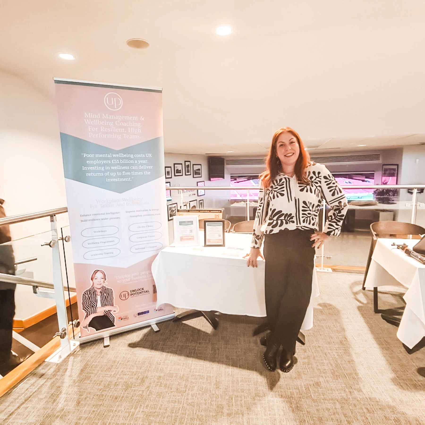 A woman stands next to a pink and light blue informational banner at a wellness or coaching event, with a table displaying framed certificates or promotional materials.