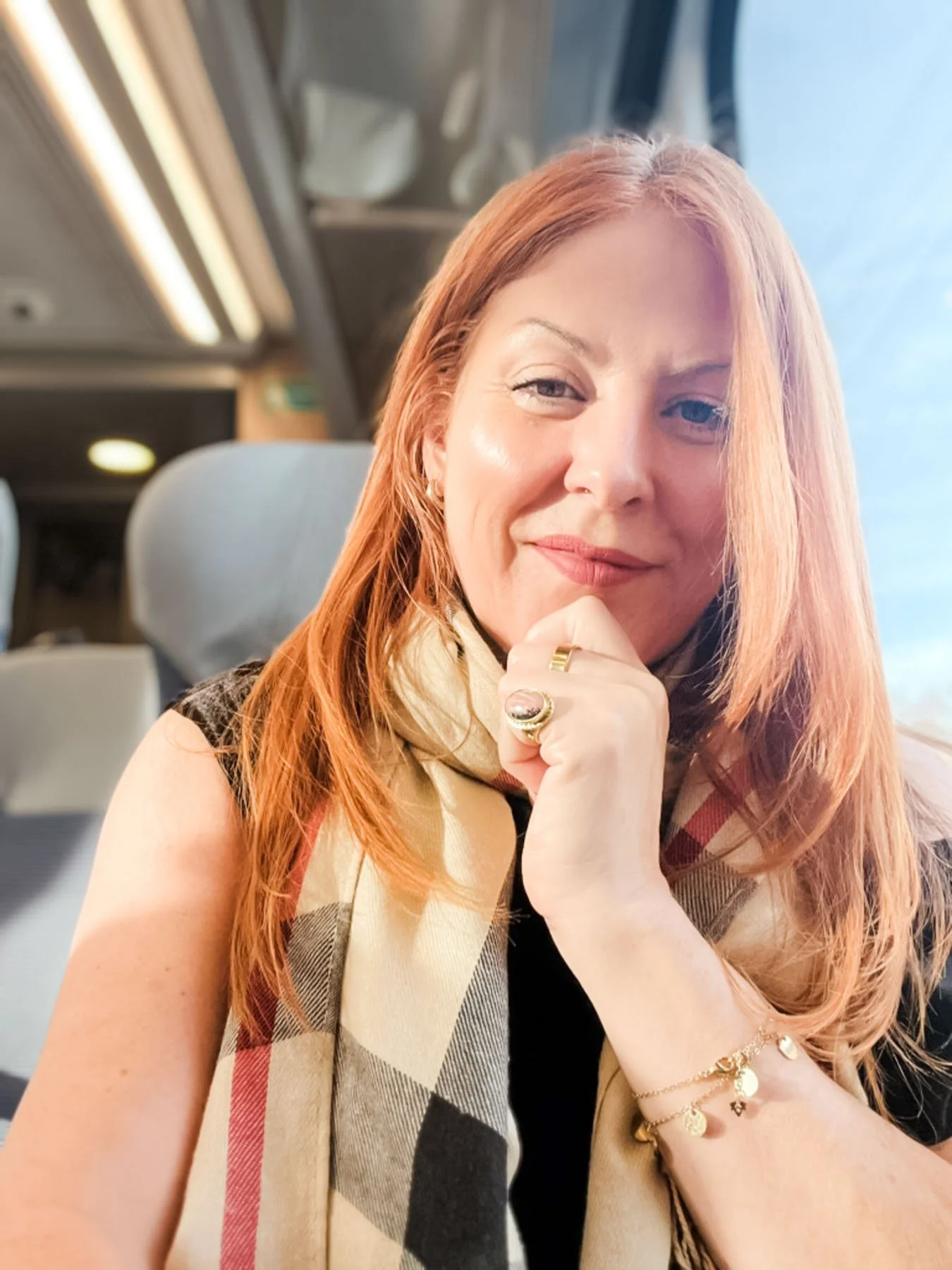 A woman with red hair smiling on a train, wearing a beige and red checkered scarf, gold jewelry, and a sleeveless black top.