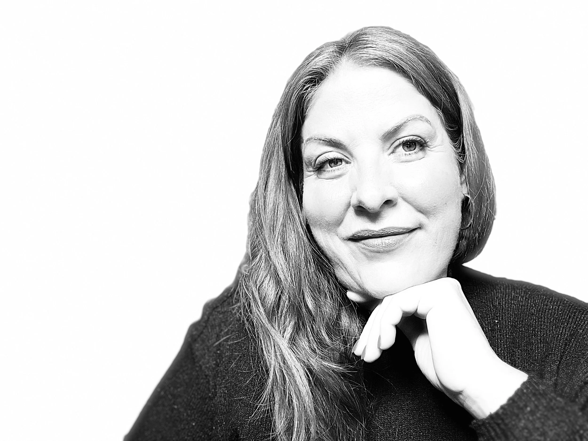 A black and white portrait of a woman with long hair, smiling gently, resting her chin on her hand, wearing a dark sweater.