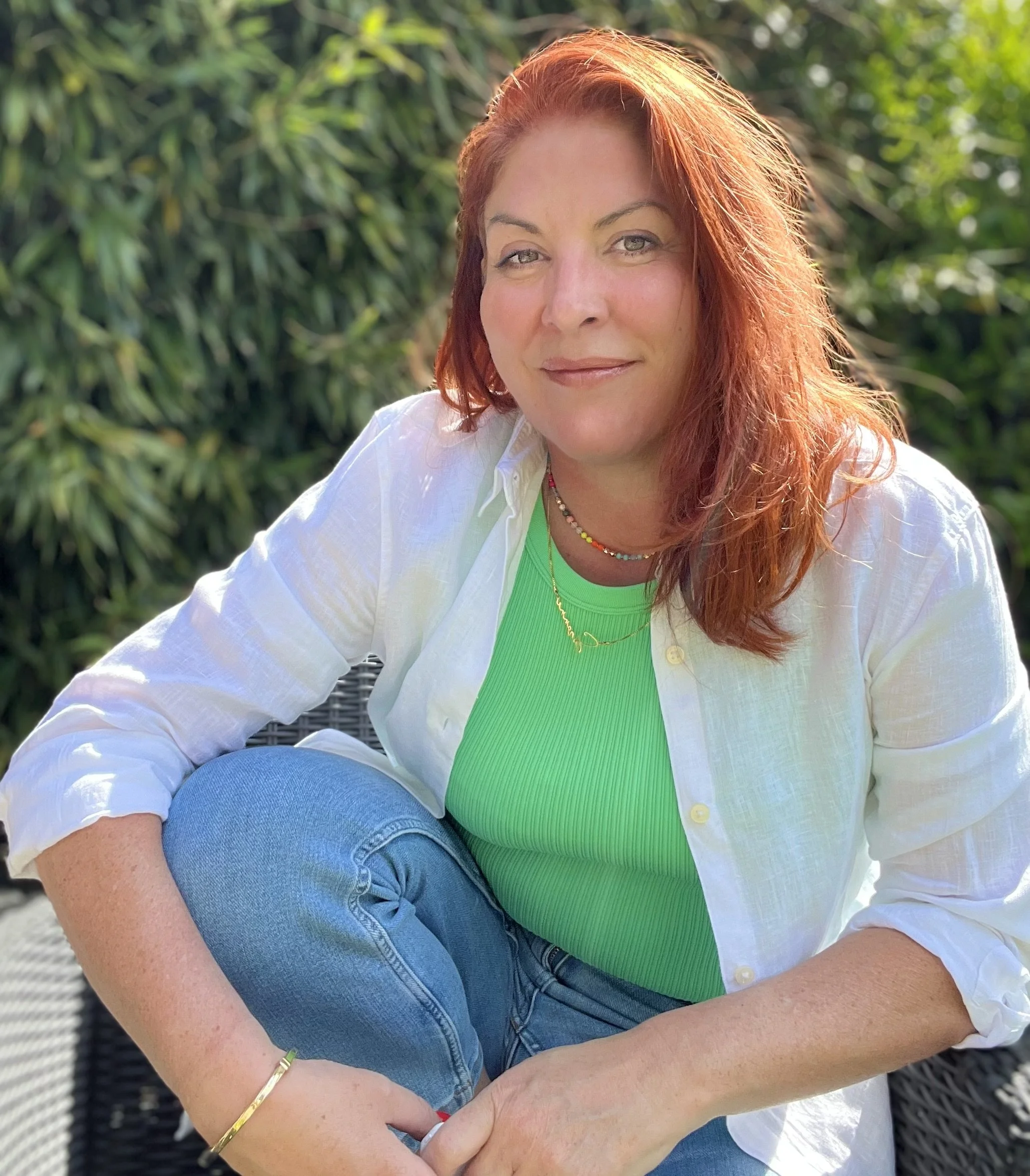 A woman with red hair sitting outdoors on a black wicker chair, wearing a white shirt over a green top, and surrounded by green foliage.