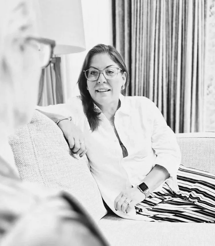A woman with glasses and a watch, sitting on a couch, smiling and engaged in conversation, in a black and white photo.