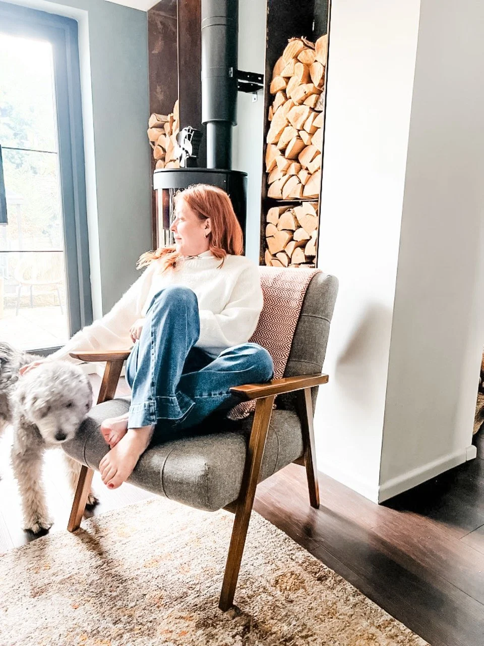 A woman with red hair sitting barefoot on a mid-century modern armchair with a dog nearby inside a cozy living room, wood logs stacked beside a black wood stove, and sunlight coming through a large window.