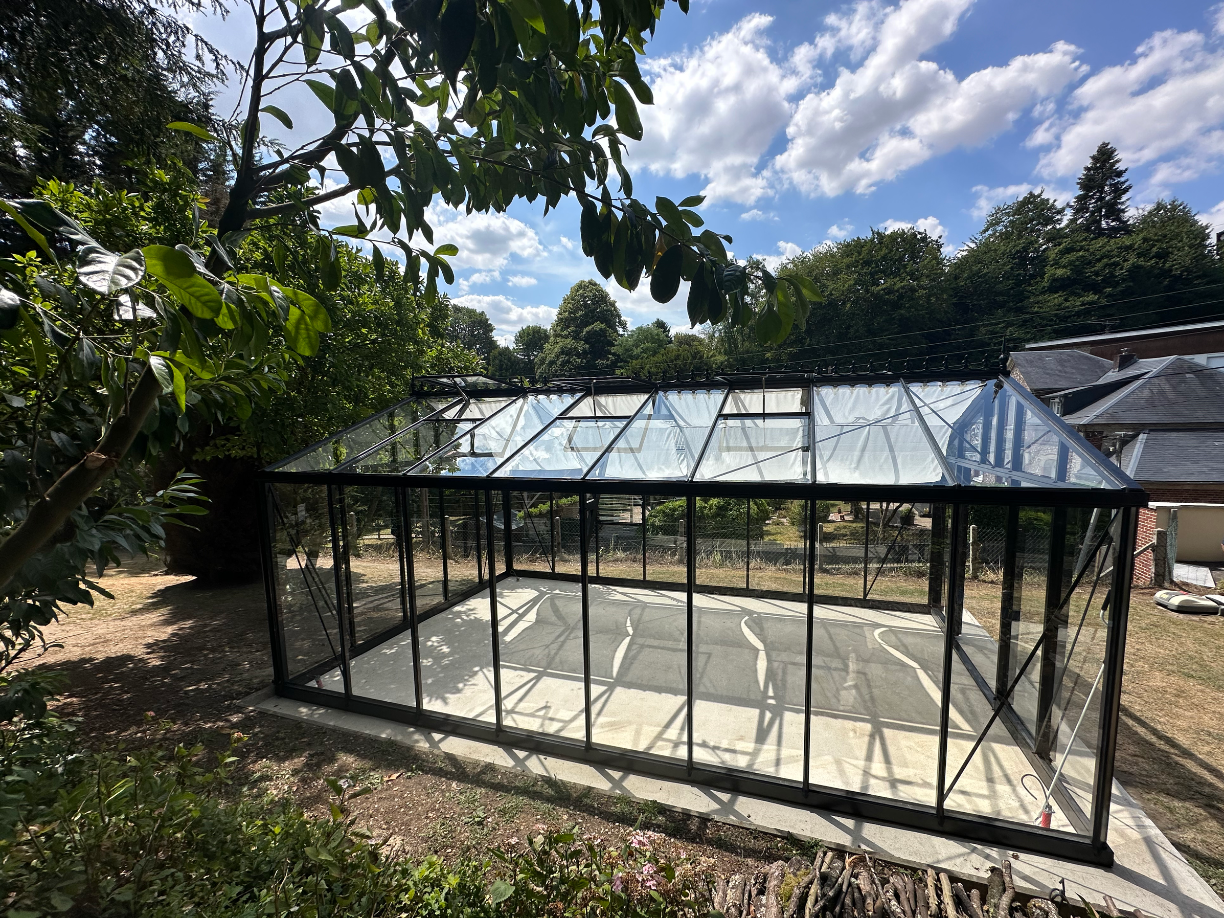 Une serre en verre située dans un jardin, entourée d'arbres verts et de ciel bleu avec des nuages blancs.