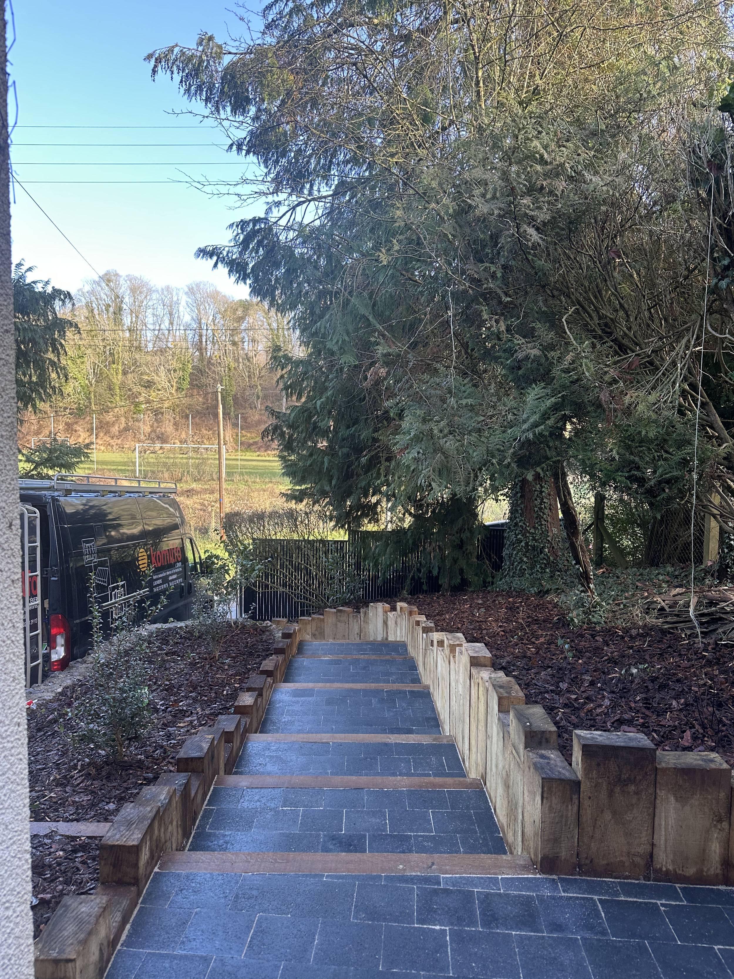Escalier en pierre avec des bordures en bois dans un jardin avec des arbres et un véhicule noir stationné à gauche.