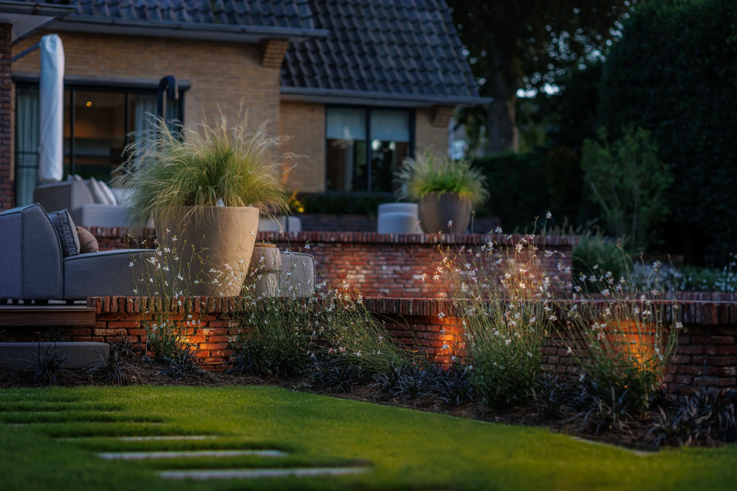 Terrasse extérieure avec plantes en pot, éclairage d'ambiance, mobilier moderne et maison en arrière-plan, during twilight.