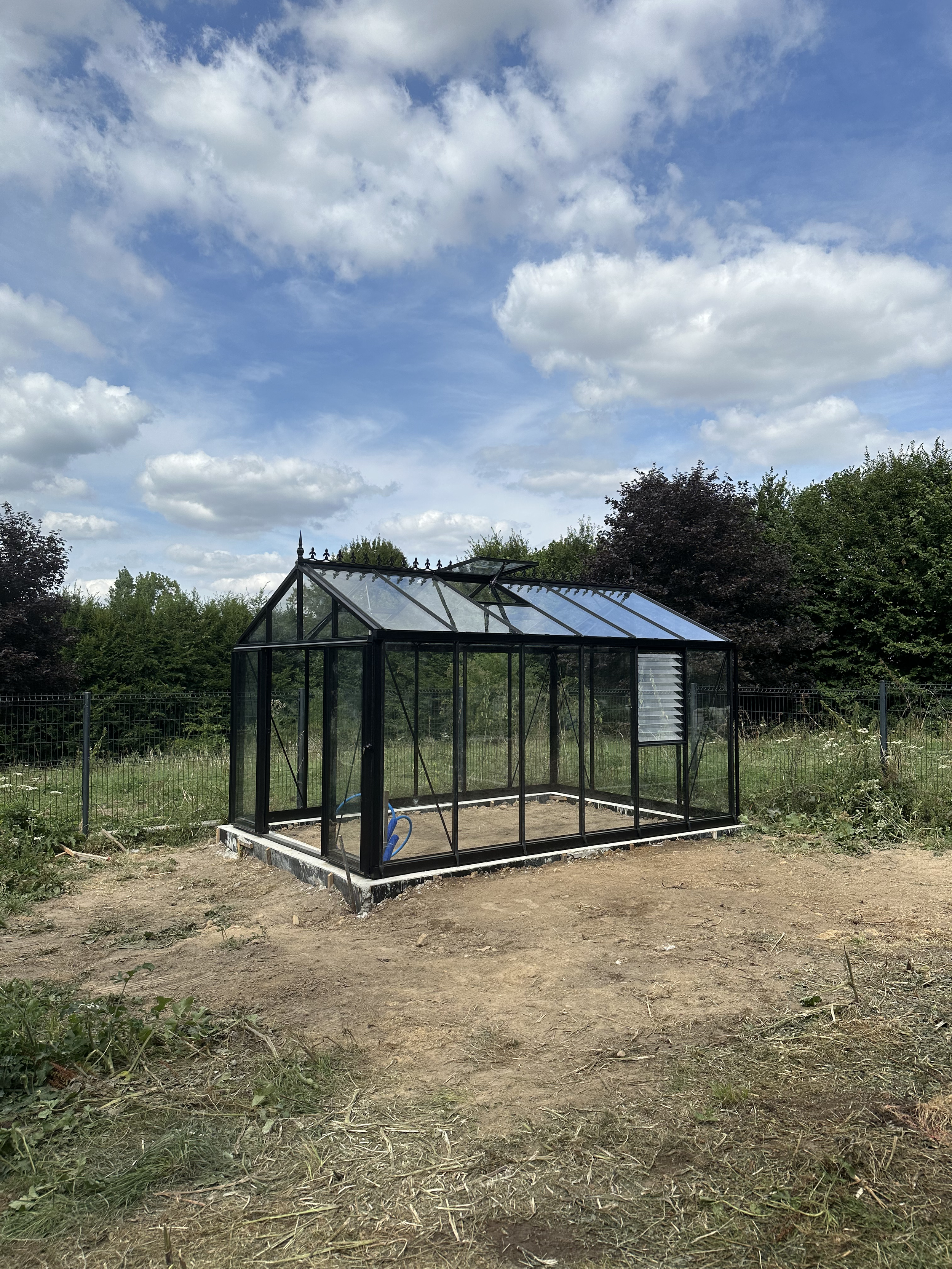 Une serre en verre noire dans un jardin entouré d'arbres et de ciel partiellement nuageux.