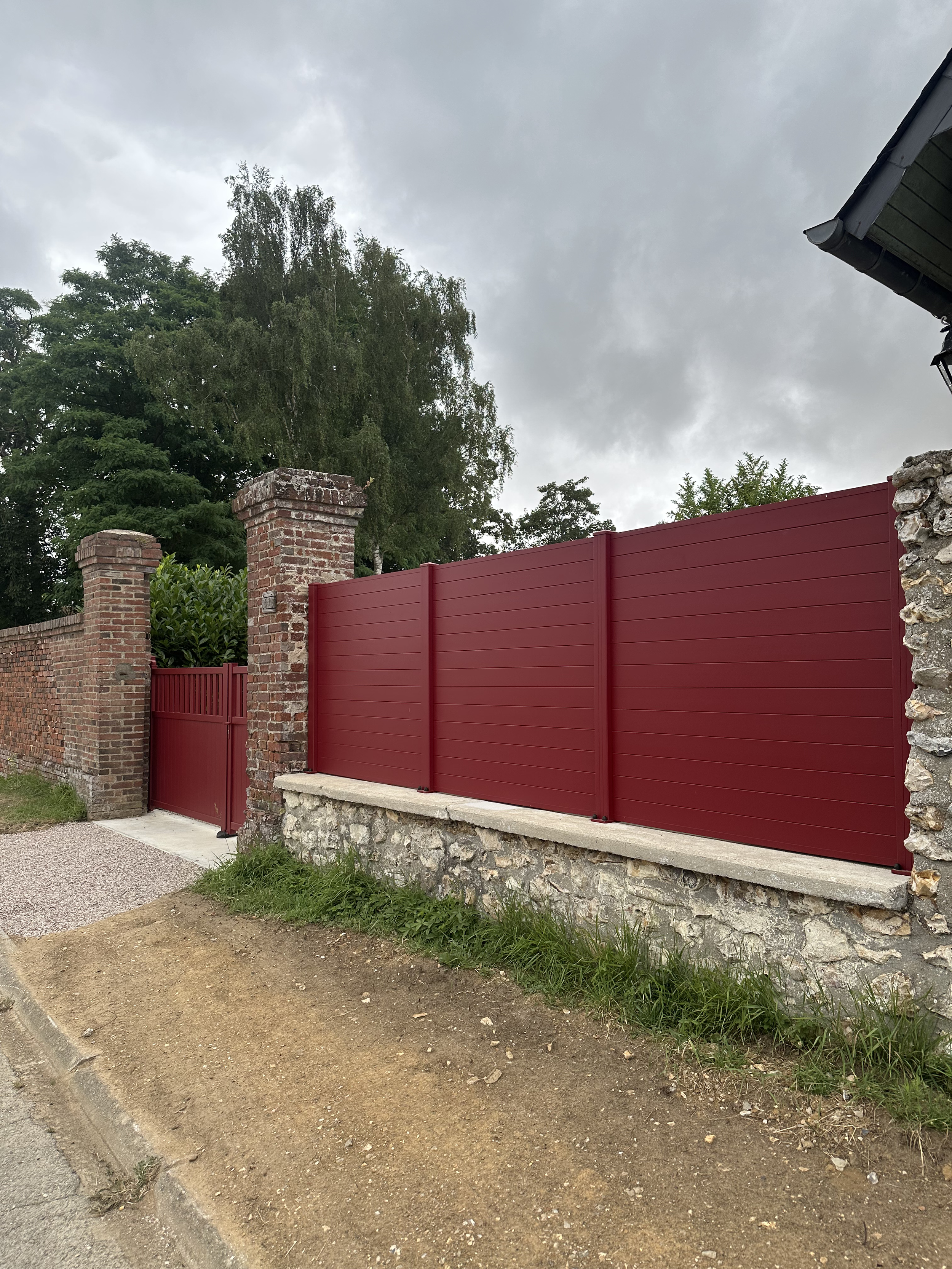 Installation d’une clôture occultante rouge bordeaux sur un muret en pierre naturelle avec piliers en briques et portillon assorti en Normandie.
