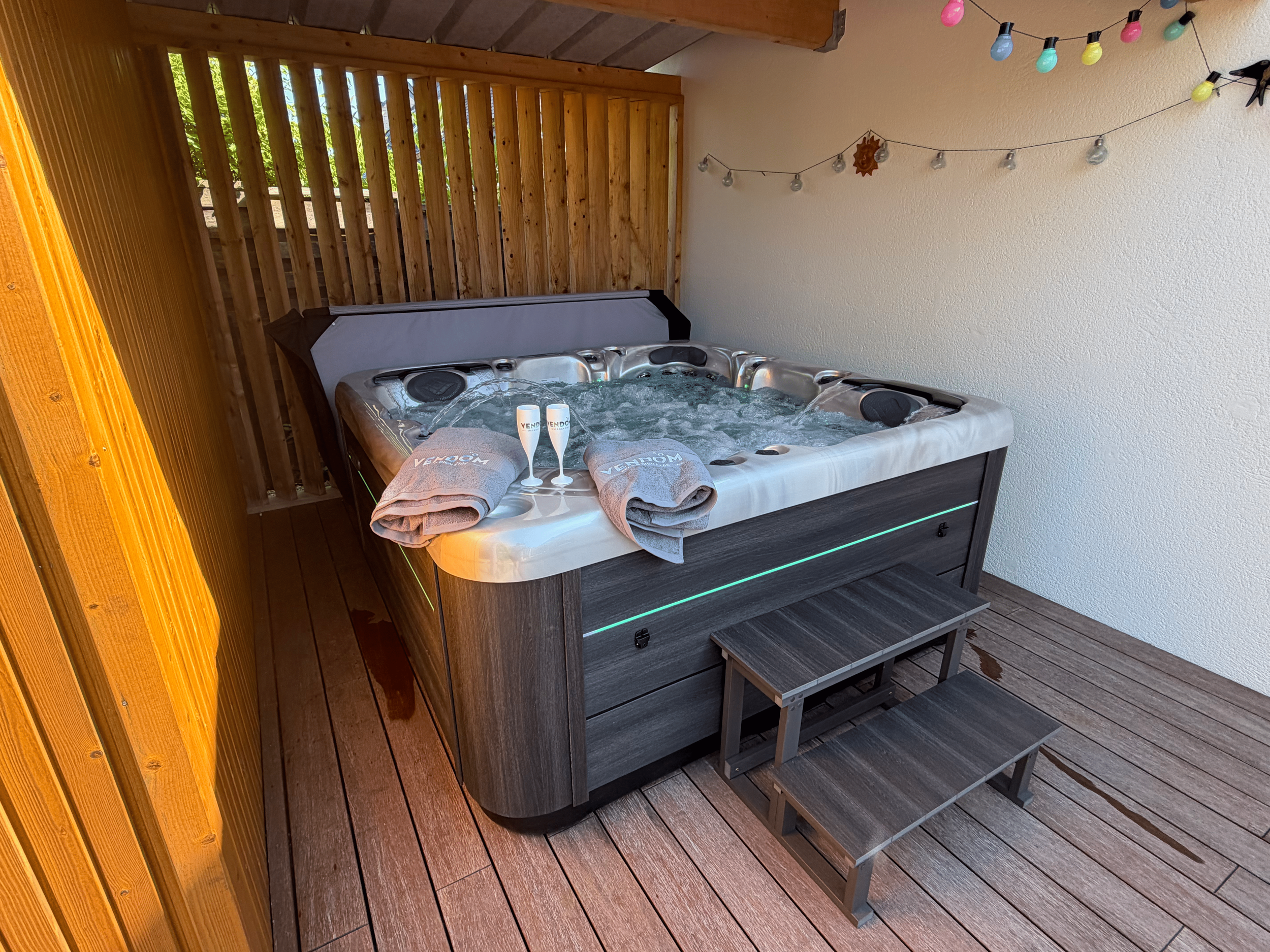 Un jacuzzi rempli d'eau avec des serviettes et des flûtes de champagne posées dessus, situé sur une terrasse en bois avec des murs en bois et des guirlandes lumineuses colorées suspendues.