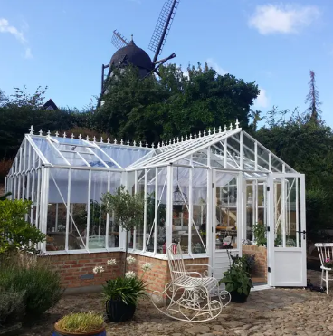 Une serre en verre avec fenêtre basculante, entourée de pots de fleurs et d'une table en bois, dans un jardin ensoleillé avec des arbres en arrière-plan.